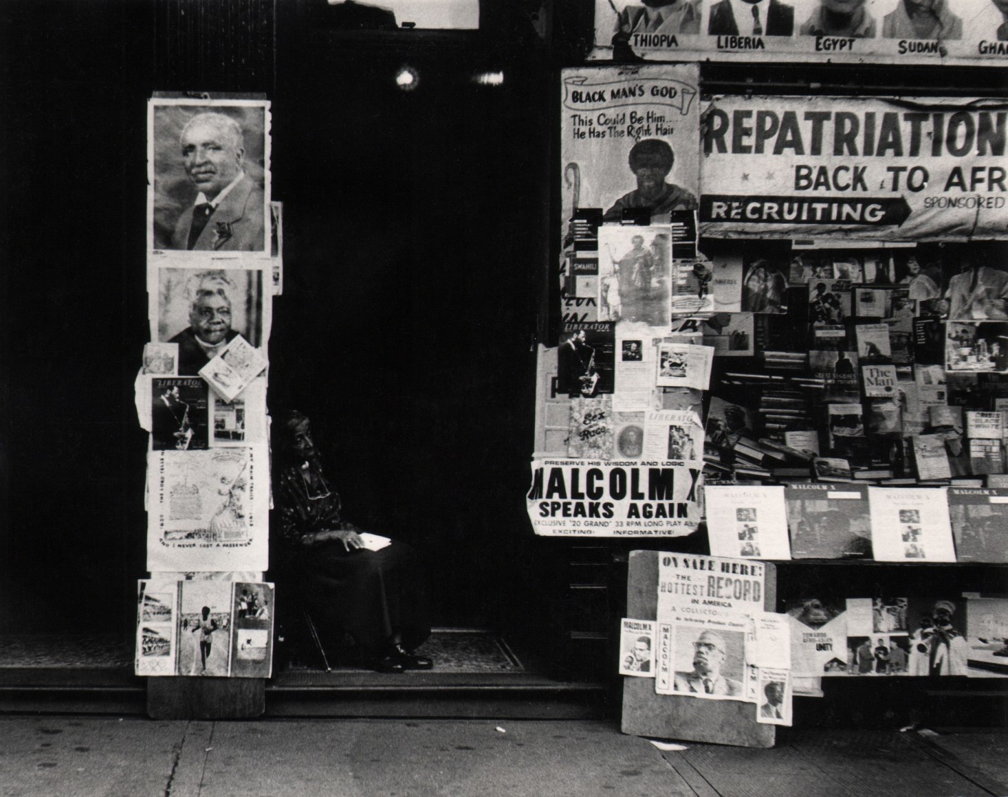 12. Beuford Smith, Woman in Doorway, Harlem, ​1965. An older woman seated in a dark doorway, surrounded by signs such as &quot;repatriation,&quot; &quot;Malcolm X,&quot; &quot;Black Man's God,&quot; etc.