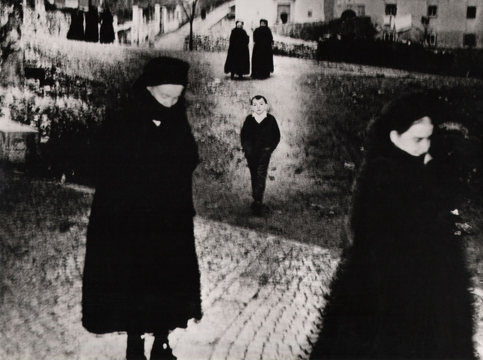 Mario Giacomelli, Scanno, ​1959/1970. High-contrast street scene with figures in black, with the central focus a young boy with hands in pockets looking to the camera.