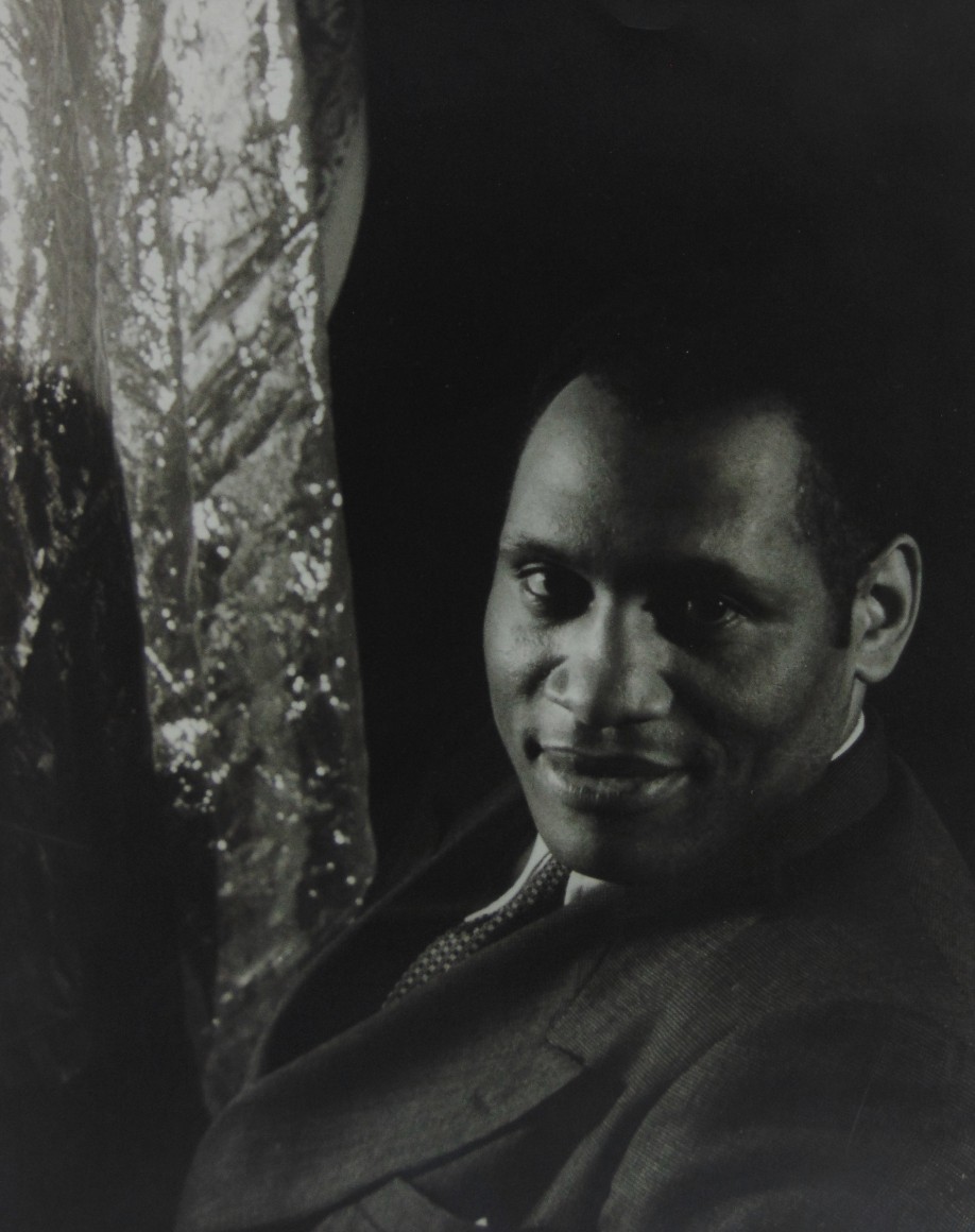 20. Carl Van Vechten, Paul Robeson, 1933. Subject smiles to the camera with torso facing left, a reflective material hangs to the left of the frame.