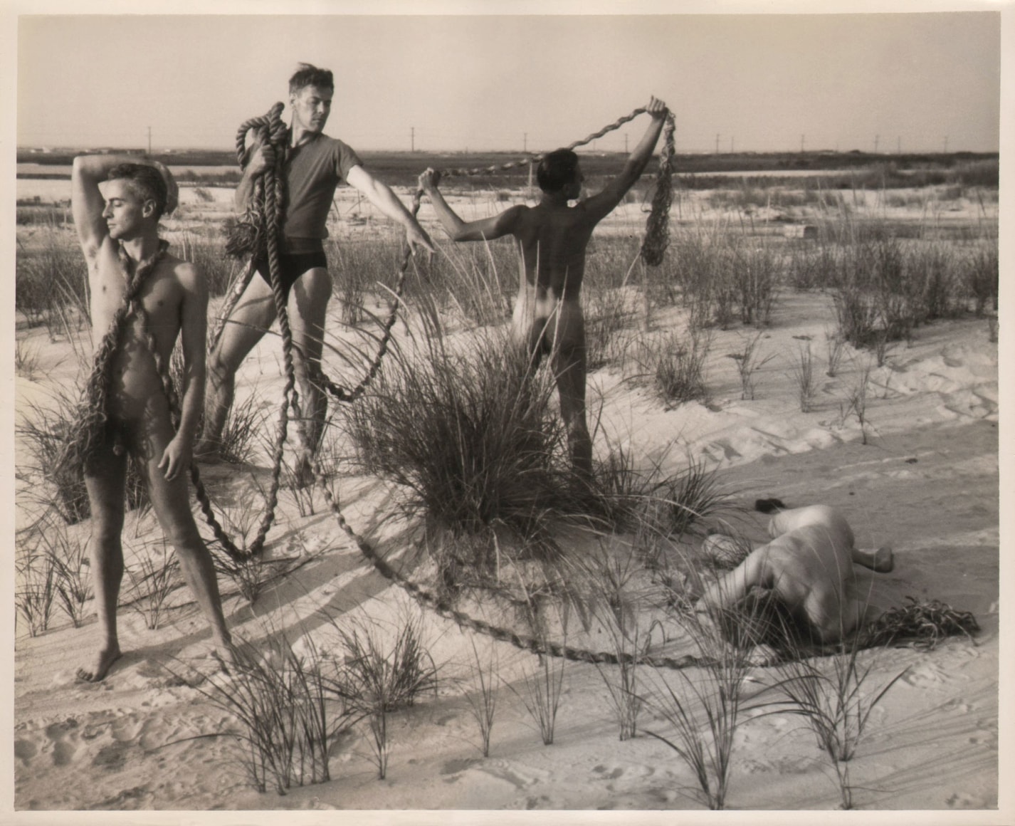 PaJaMa, Glenway Wescott, George Platt Lynes, Paul Cadmus, [unidentified], ​c. 1941. Four men on the beach with a thick rope connecting them. Three are standing (left), one lays facedown in the sand.