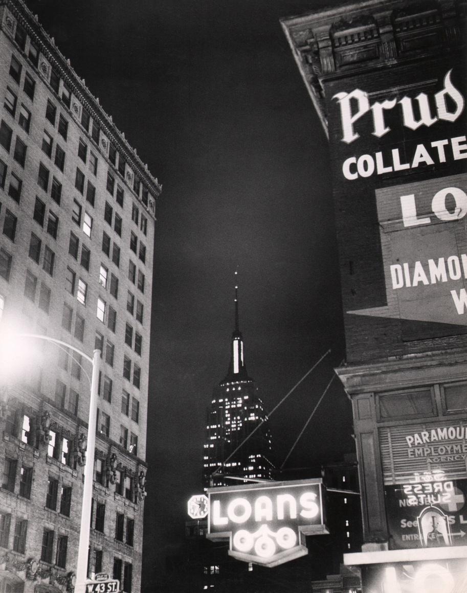 34. Weegee, West 43rd Street, ​c. 1950. Nighttime street view facing the Empire State Building pictured between an unmarked building on the left and a &quot;Loans&quot; business with a neon sign on the right.