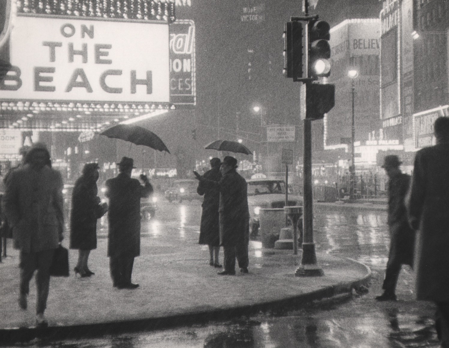 49. Bedrich Grunzweig, Times Square at Night, New York City, ​c. 1959. Pedestrians standing on a street corner, some holding umbrellas. Nearby is a marquee that reads &quot;On the Beach&quot;.