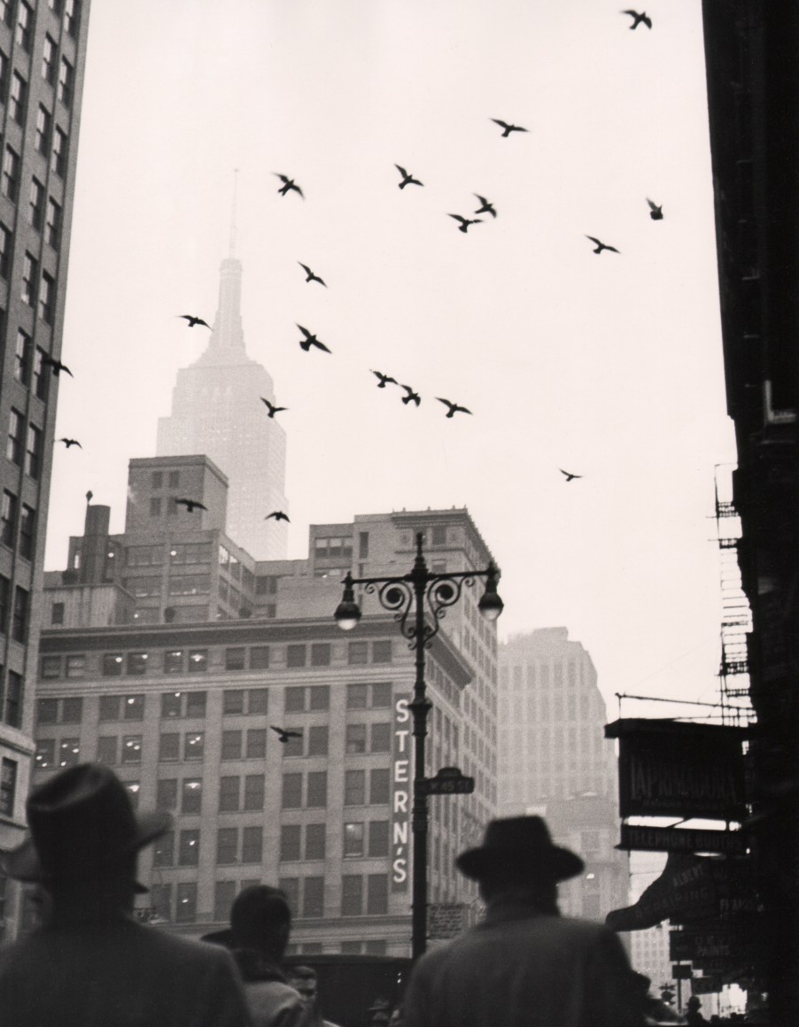 39. Benn Mitchell, 6th Avenue, NYC, ​1949. Vertical street view featuring men in hats in the foreground, a building marked &quot;Stein's&quot; and flying birds in the midground, and a fog-obscured Empire State Building in the left background.