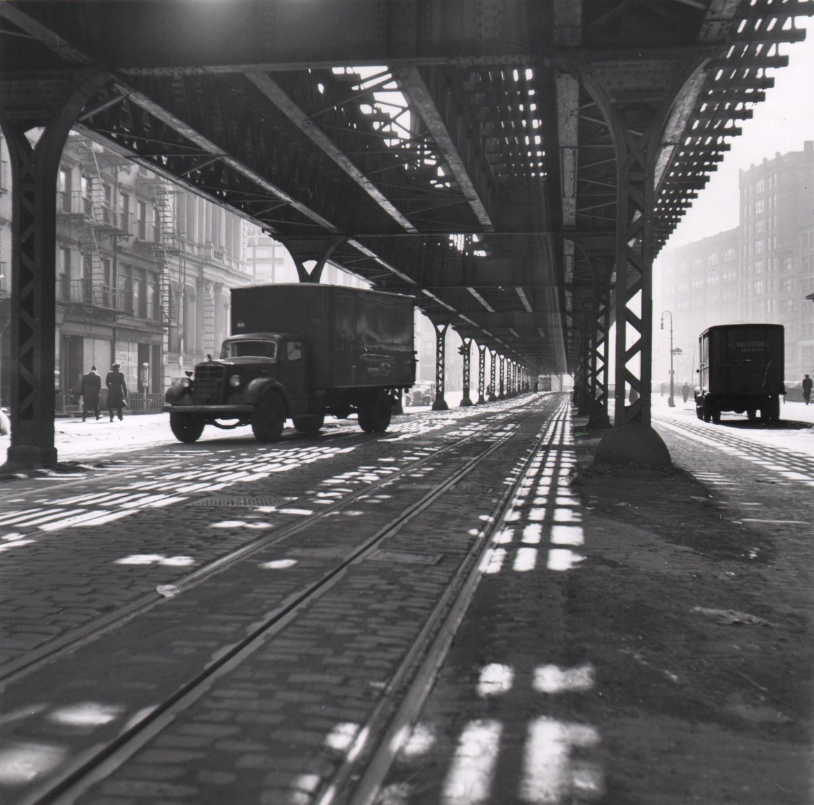 1B. Louise Rosskam, New York City: Trucking. Trucks under the Third Avenue elevated platform, 1945. Two trucks beneath an elevated train platform with daylight streaming down to the ground beneath.