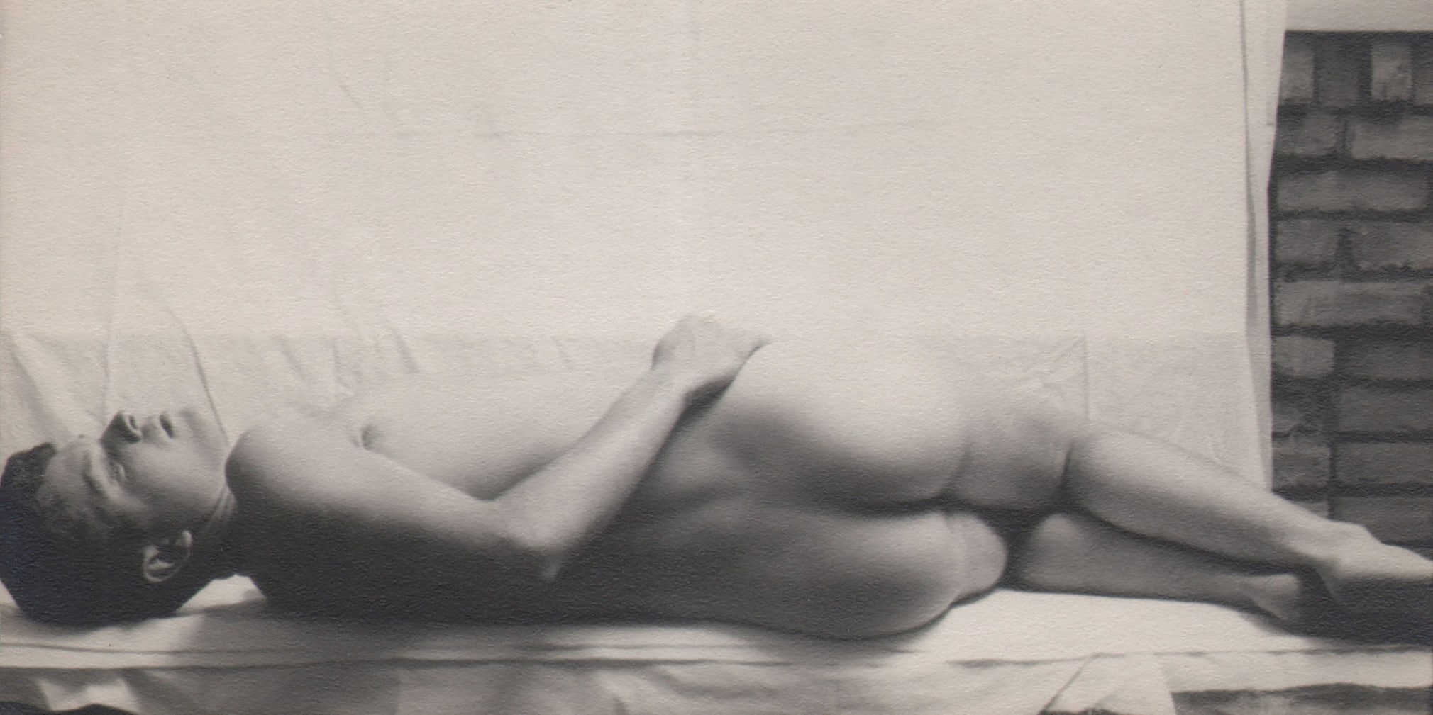 PaJaMa, Sandy Campbell, ​1943. Reclining male nude filling the lower portion of the horizontal frame against a white fabric backdrop. He lays on his side away from the camera, head turned upward.