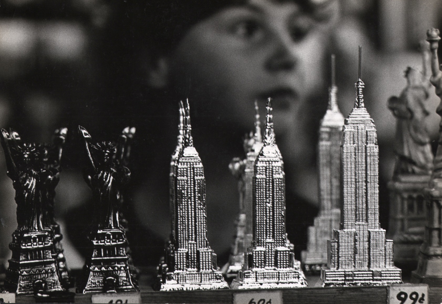 09. Jan Lukas, Souvenirs of New York, ​1964. Miniatures of the Statue of Liberty, Chrysler Building, and Empire State Building for sale on a shelf. A child's face is out of focus behind them.