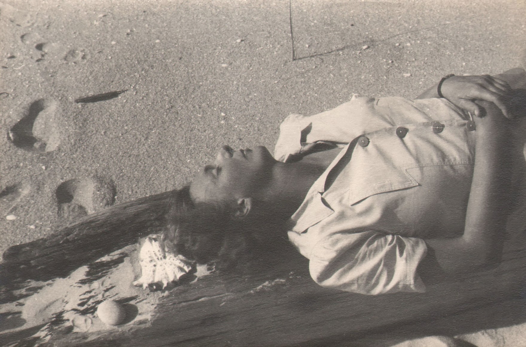PaJaMa, Margaret French, Fire Island, c. 1945. A woman in white's upper torso extends from the right of the frame; she is laying on a piece of driftwood on the beach.