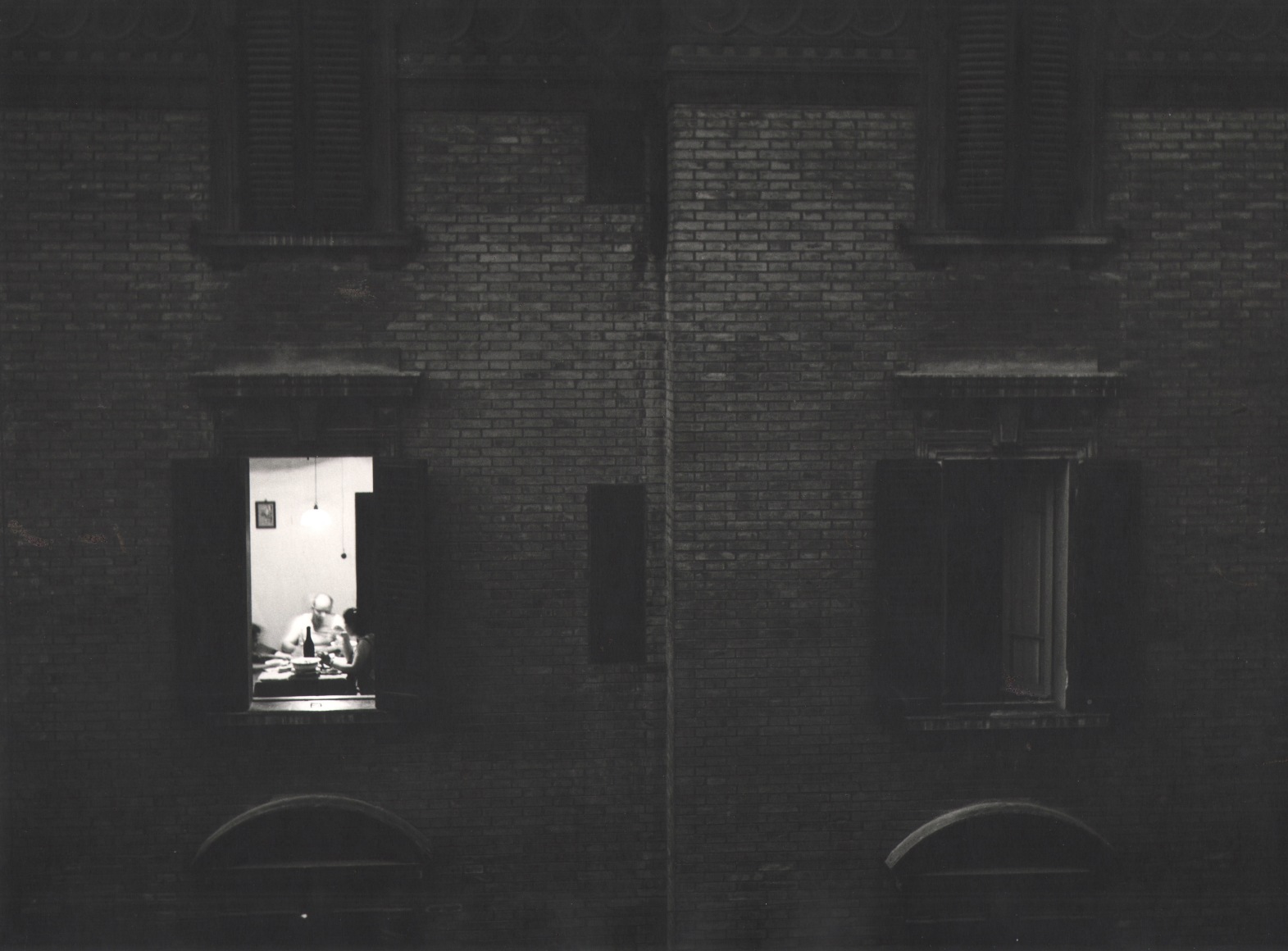 Nino Migliori, Summer evening from 'People of Emilia,' 1953. Exterior view of an apartment building. One window is lit and a family is seated at the dinner table.