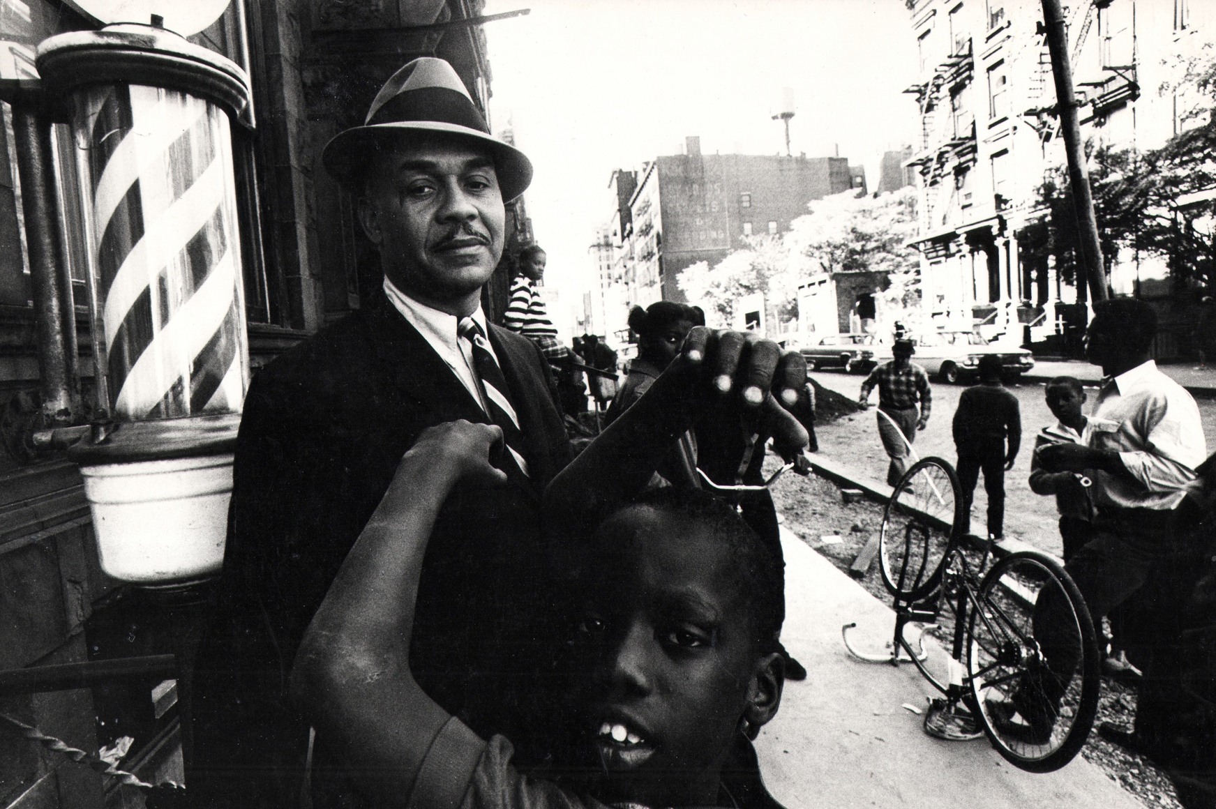 David Attie, Ralph Ellison in Harlem, ​1966. Subject poses on the street beside a barber shop, looking into the camera as various figures move around him.