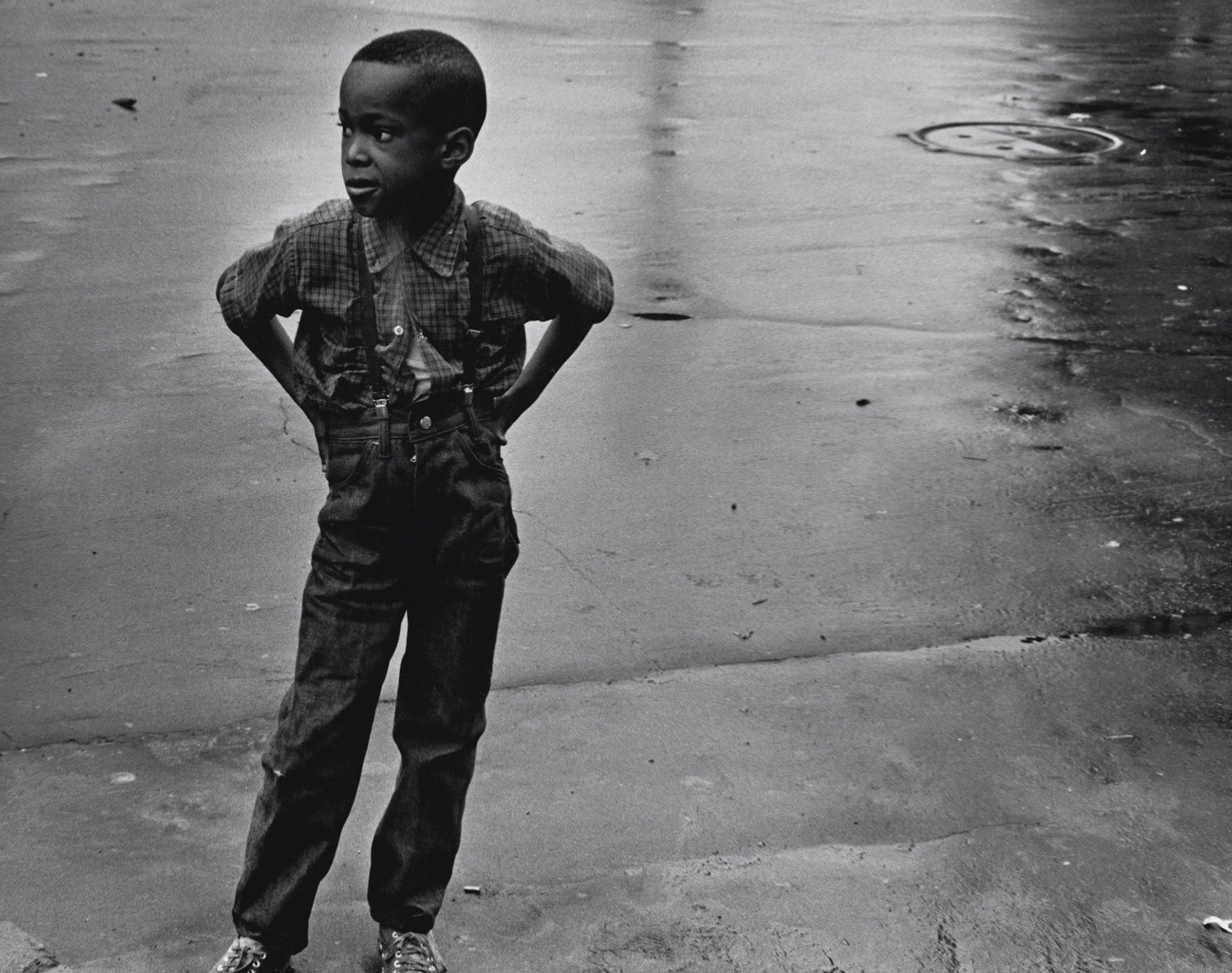 22. Beuford Smith, Boy in Street, Brooklyn, ​1969. A young boy stands on a wet street with hands on hips, looking to the left of the frame.