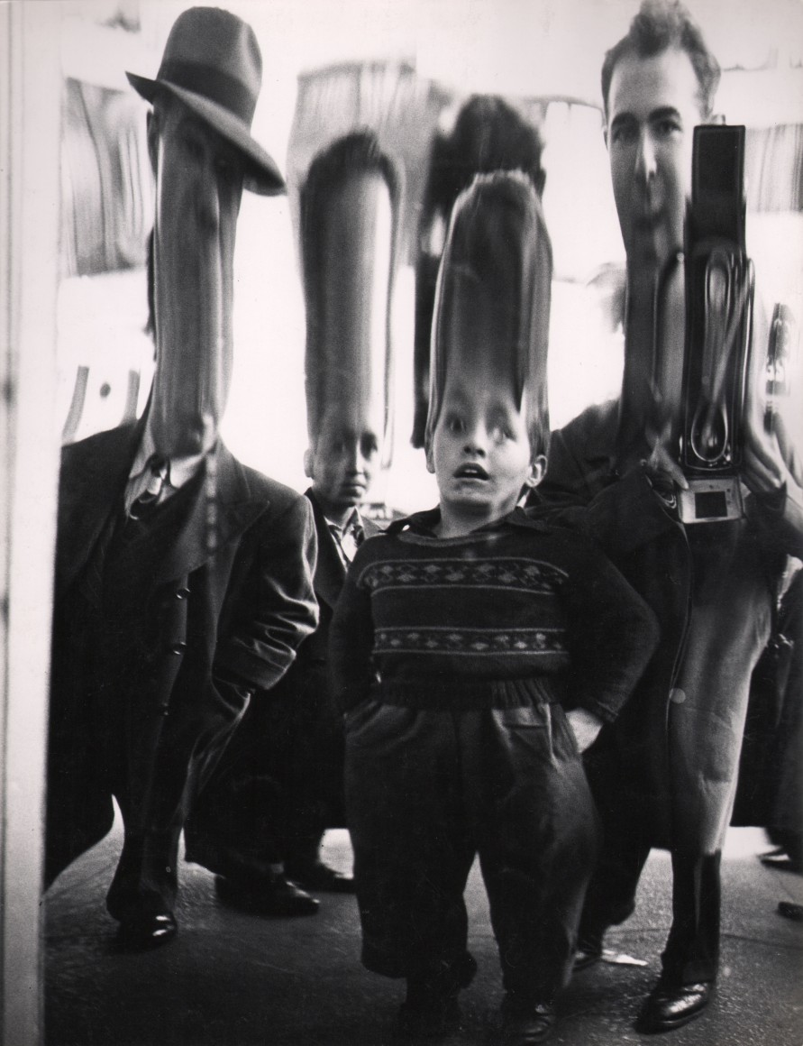 37. Benn Mitchell, Mirrors of Life, Self-Portrait, 42nd Street, ​1950. Funhouse mirror-like distortion of the photographer and three other figures in which their heads are vertically stretched.