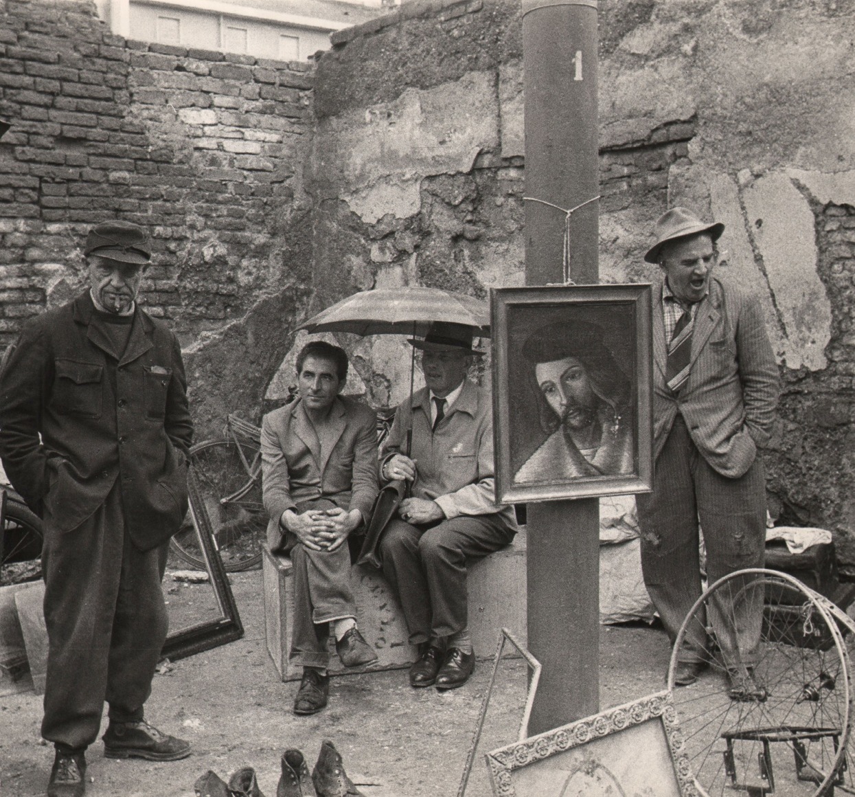 Mario Finocchiaro, Milano - Fiera di Sinigaglia, ​1959. Two men seated beneath a small umbrella are flanked by two men standing amongst various objects in a yard sale-like setting.