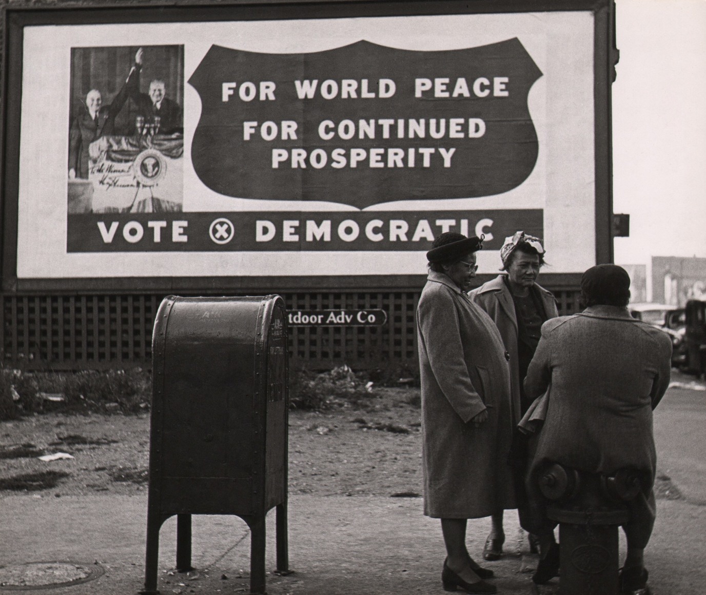 Marvin E. Newman, Chicago, ​1950. Three adults chatting by a mailbox, one seated on a fire hydrant. A billboard in the background reads &quot;For world peace, for continued prosperity, vote Democratic