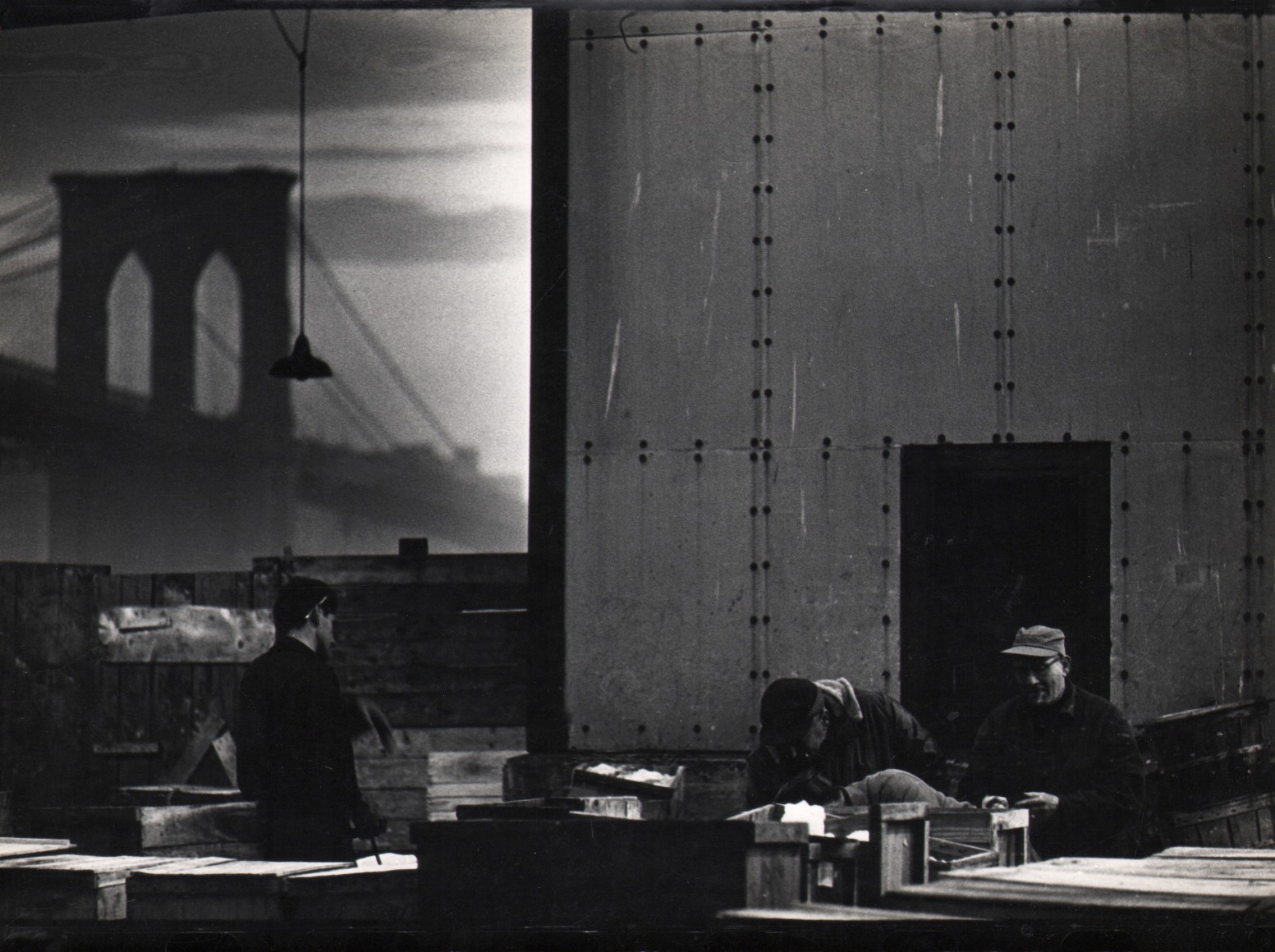 12. Jan Lukas, New York, Fulton Fishmarket, ​1964. Three men standing in front of a metal structure. The Brooklyn Bridge is seen in the background left.