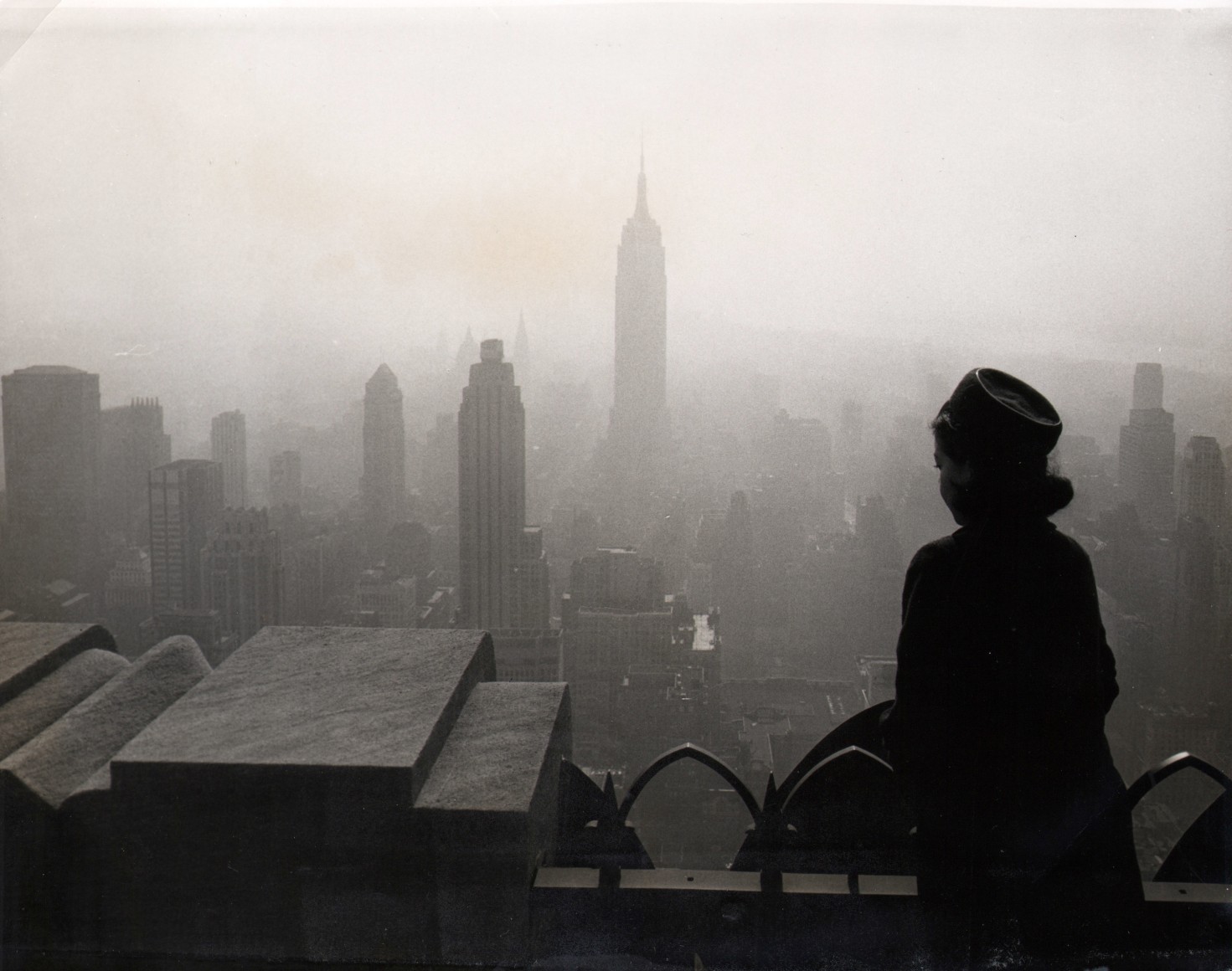 17. UPI Photo, Smog-Bound City, ​1965. A silhouetted woman stands on a balcony, looking out towards a foggy skyline that includes the Empire State Building at the center.