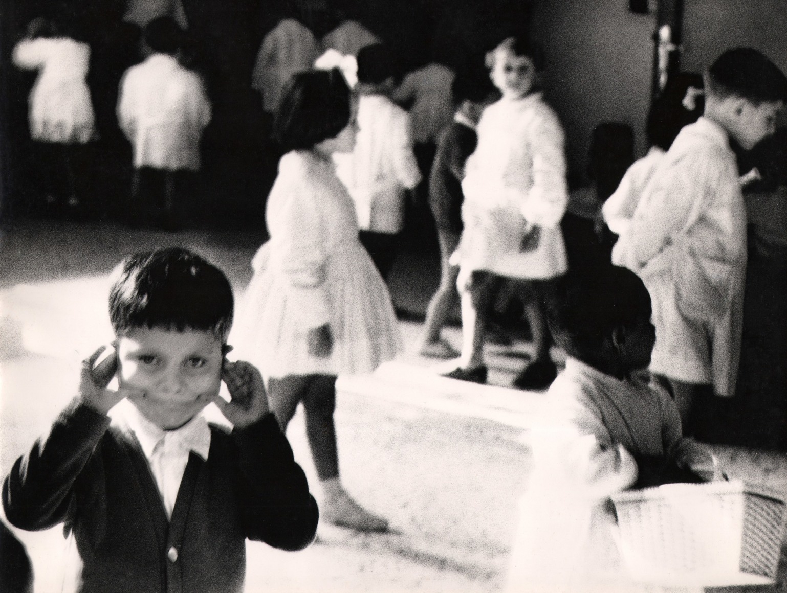 17. Renzo Tortelli, Piccolo Mondo, 1958&ndash;1959. High contrast image. A small boy in the lower left of the frame distorts his face using his hands. Various out-of-focus children play in the background.