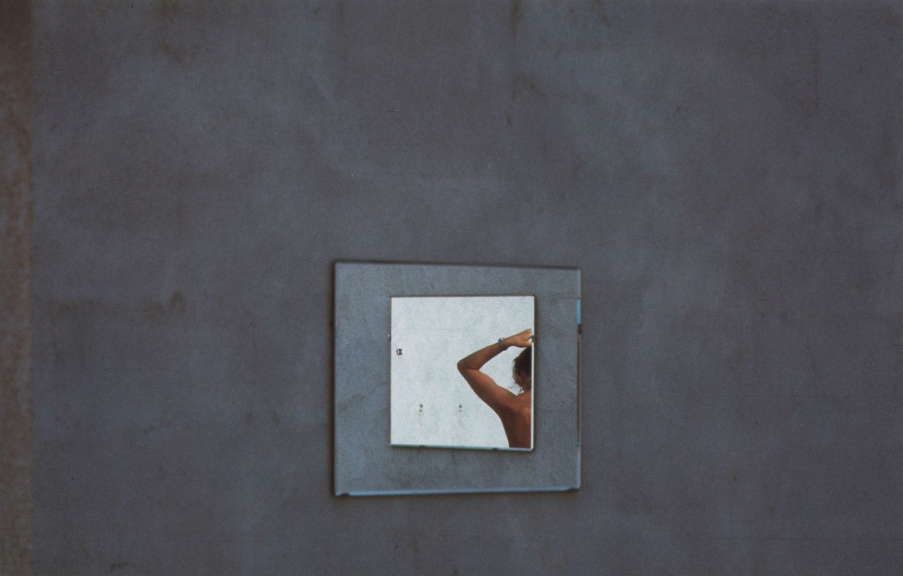 Luigi Ghirri, Untitled, ​1976. Color photograph of a blue wall with two concentric squares in the center. The innermost shows the left arm, upper back, and head of a nude figure facing away, reaching upward to their hair.