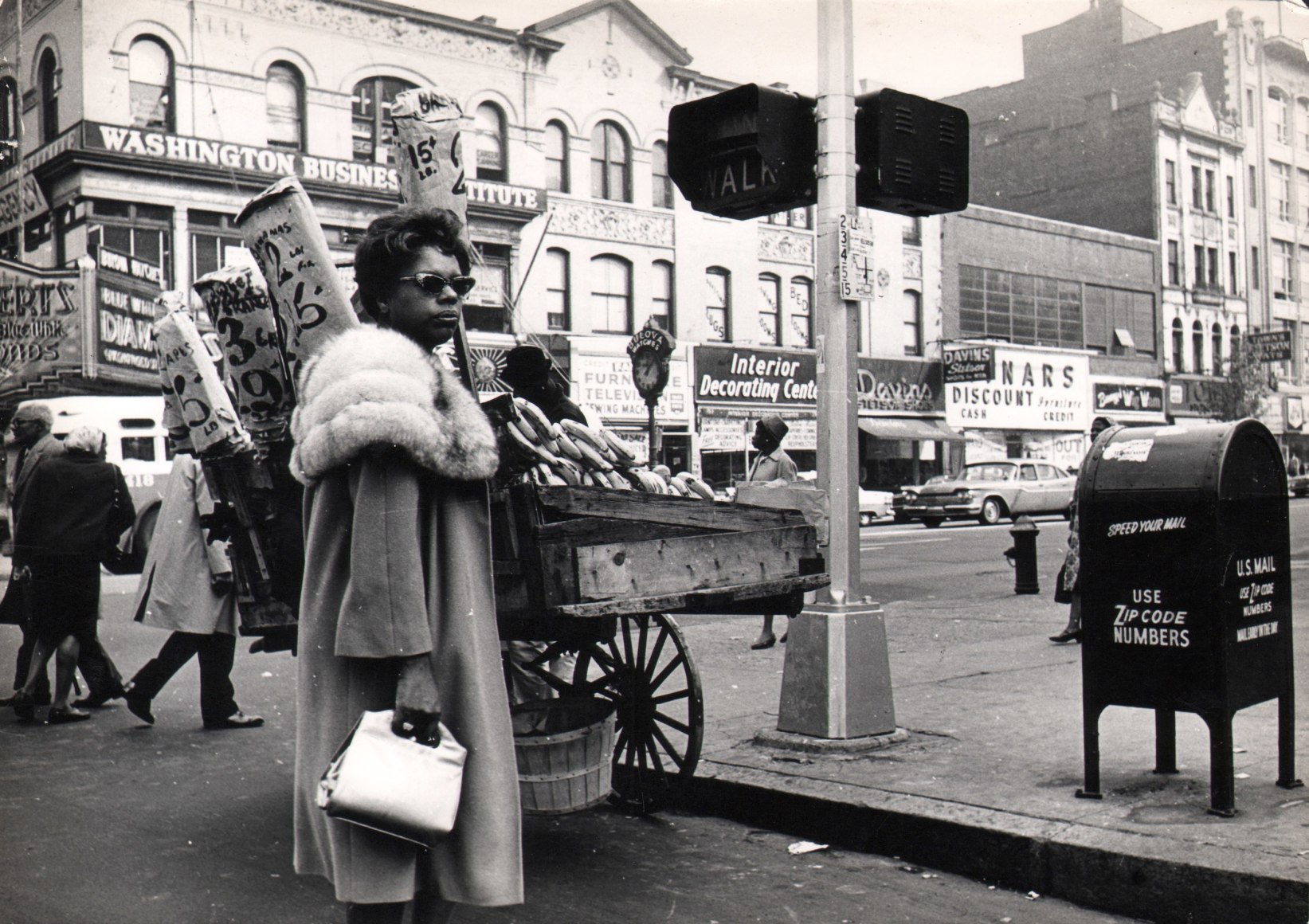 10B. Jan Lukas, Untitled, 1964. A woman in sunglasses and fur stands in the foreground at a street corner in front of a produce cart.