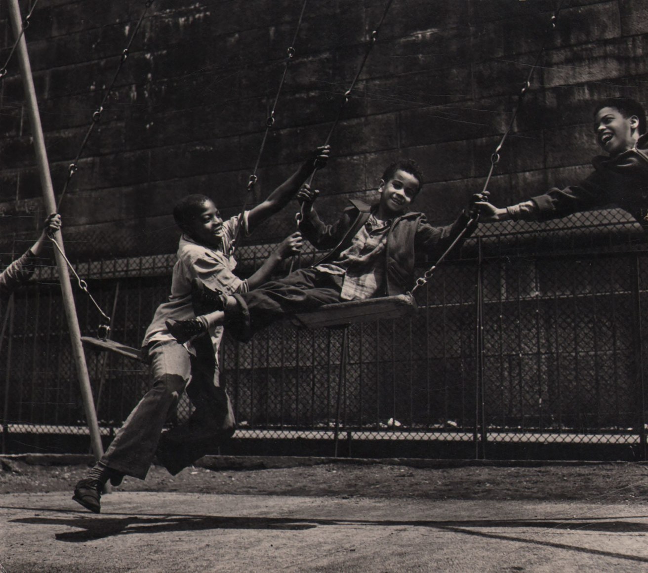 Ken Heyman, Young Crucifix, ​c. 1963. Three young boys play on a swing set; the boys to the left and right reach to hold onto the center swing.