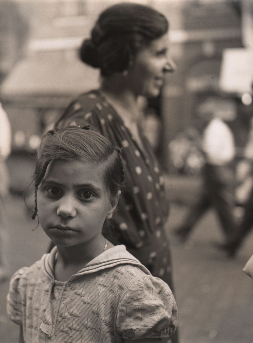 Clemens Kalischer, New York Street Scene, Italians, c. 1955&ndash;1960. A young girl looks to the camera in the foreground while a woman stands, facing right, out of focus behind her.