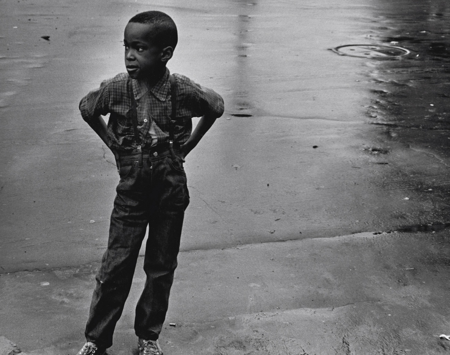 29. Beuford Smith, Boy in Street, Brooklyn, 1969. A young boy in suspenders stand with hands on hips on wet pavement, looking to the left of the frame.