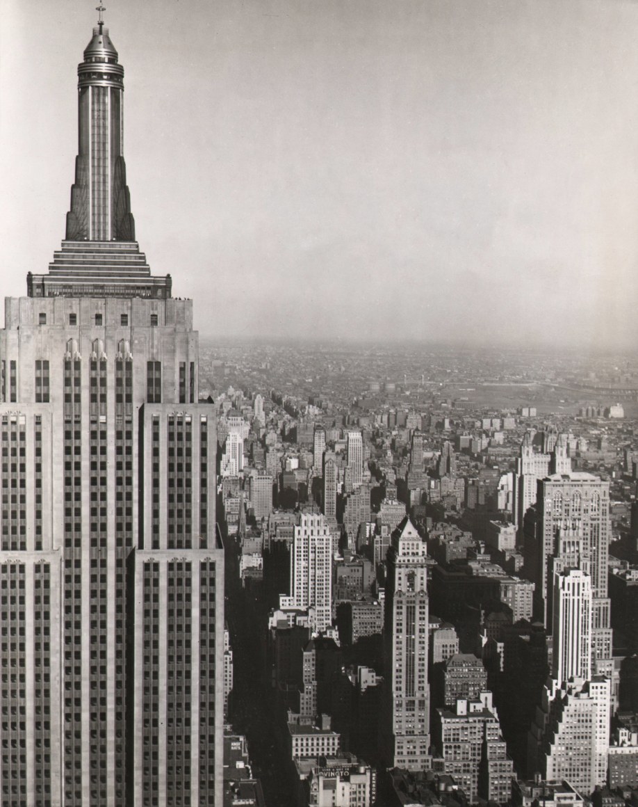 21. Anonymous, Heading Straight for the Empire State Building, 1939. Straight- on view of the top half of the Empire State Building, about level with the highest observation deck. The building occupies the left third of the frame, with the city skyline and horizon on the right.