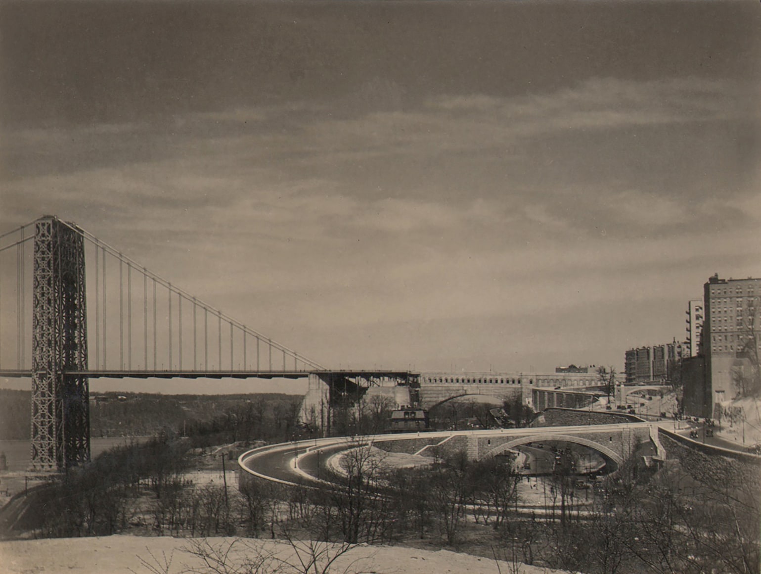 Paul J. Woolf, George Washington Bridge, c. 1933. Cityscape with bridge on center left of the frame. Cloudy sky above. A few buildings are visible on the far right.