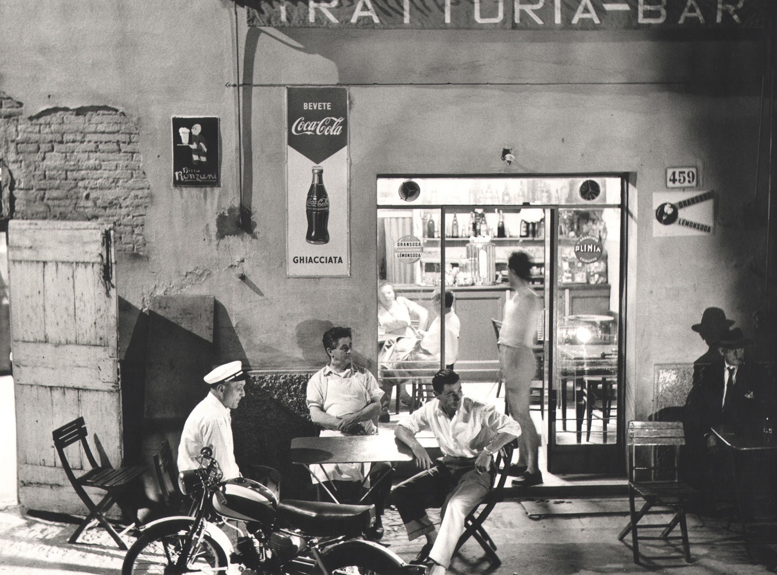 Nino Migliori, Gente dell'Emilia, ​1959. Men seated at a table outside a building marked &quot;Trattoria Bar&quot;. Soda advertisements appear on the walls and windows.