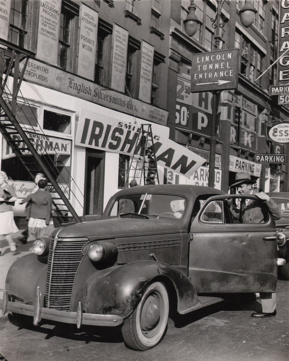 7B. Weegee, Untitled, ​c. 1945. A car in the foreground beneath a &quot;Lincoln Tunnel Entrance&quot; sign. Men on a ladder hang a sign that reads &quot;Smiling Irishman&quot; on a storefront in the background.