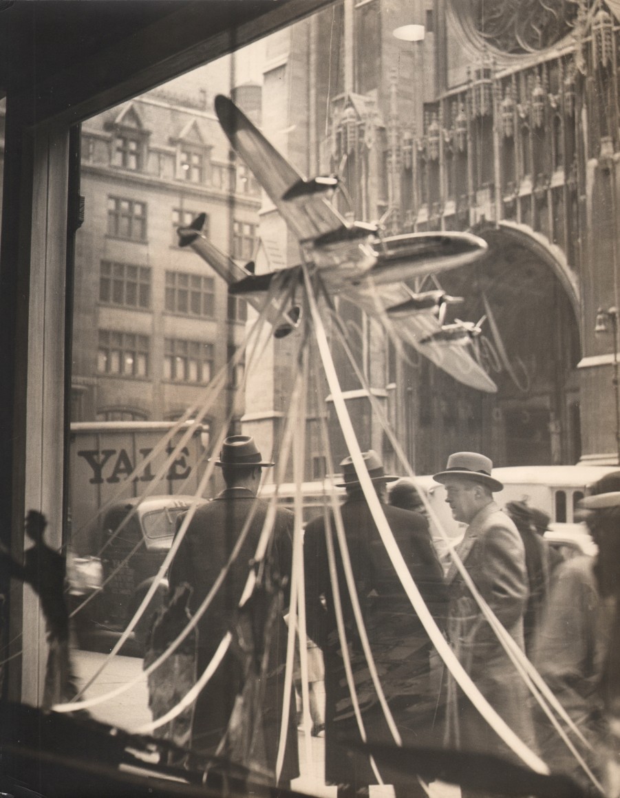 5B. Simpson Kalisher, Untitled, ​c. 1948. View out a shop window with a model plane hung in the foreground. Men gather on the sidewalk outside across from a cathedral.