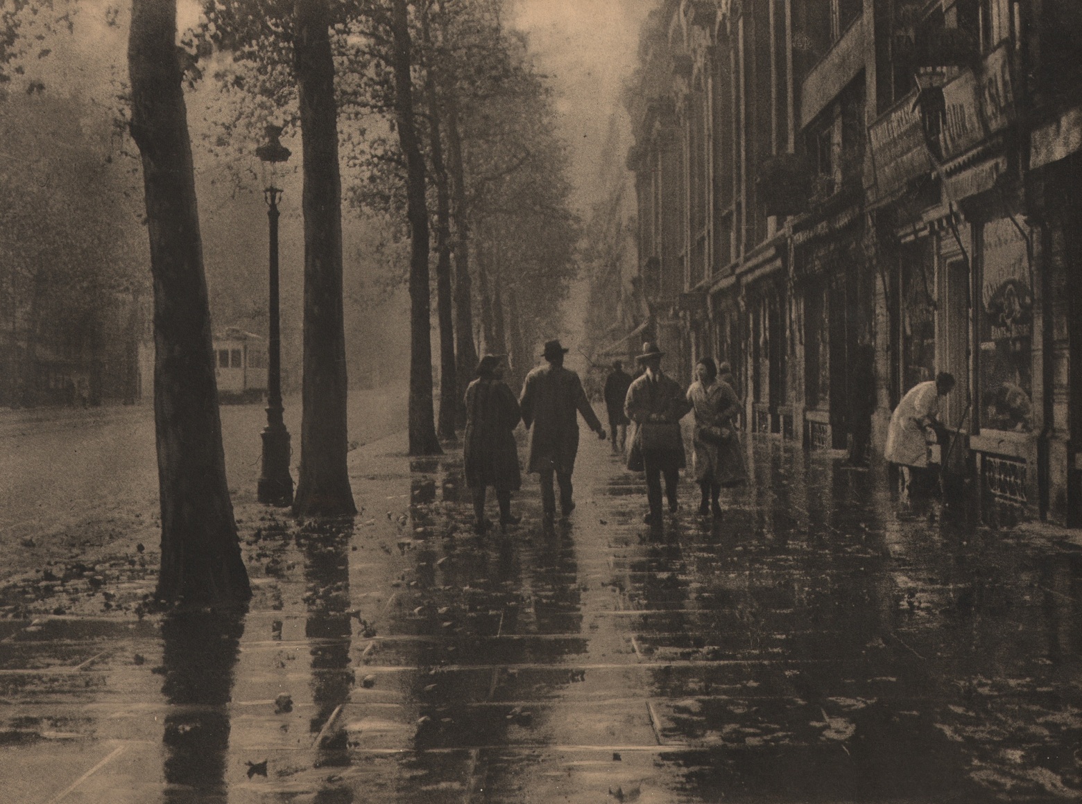 02. L&eacute;onard Misonne, Untitled, c. 1932. Pedestrians walk a tree-lined, wet sidewalk. Sepia-toned print.
