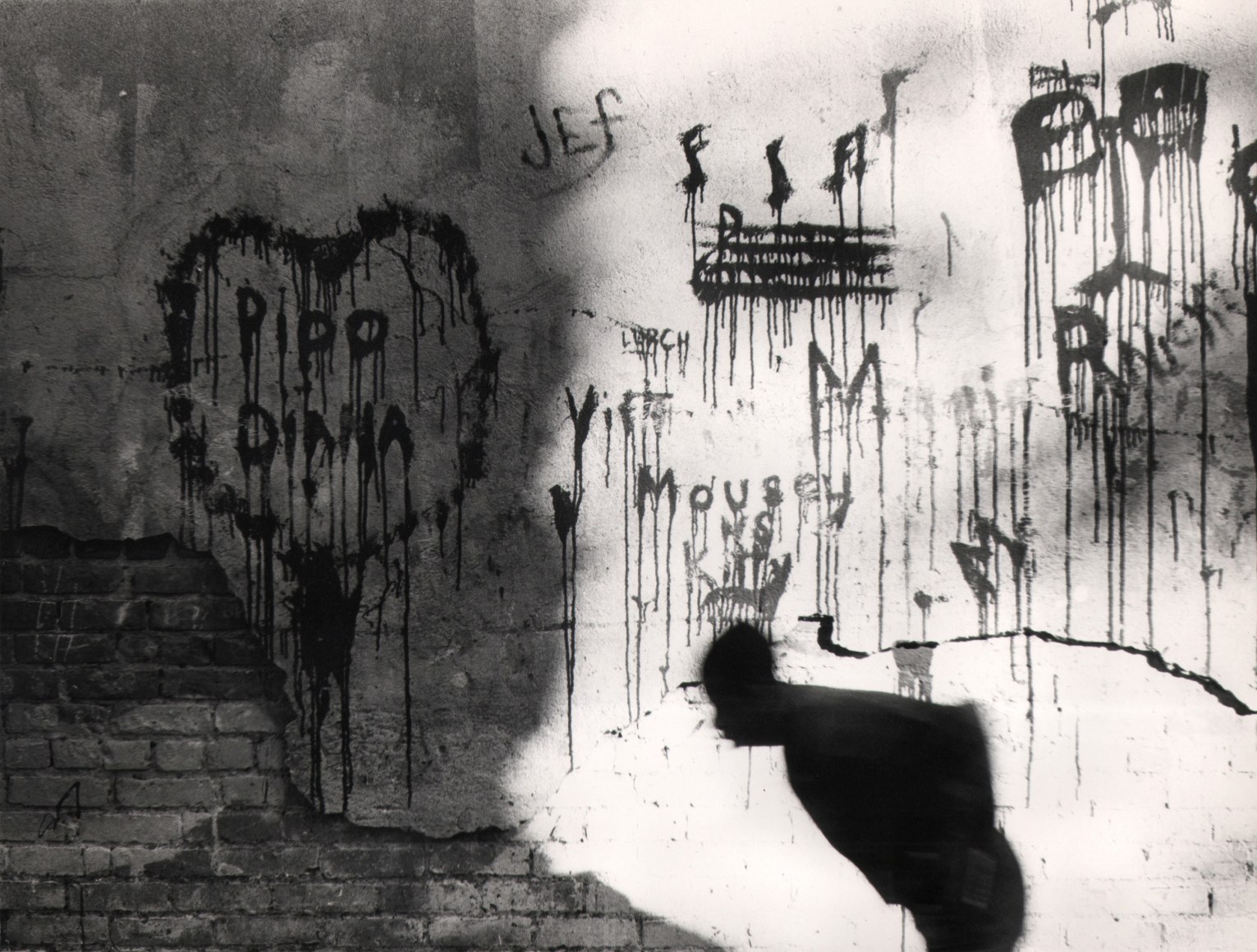 21. Beuford Smith, Wall, Lower East Side, ​1972. A blurred, hunched silhouette moves across the frame towards the left against a wall covered in graffiti.