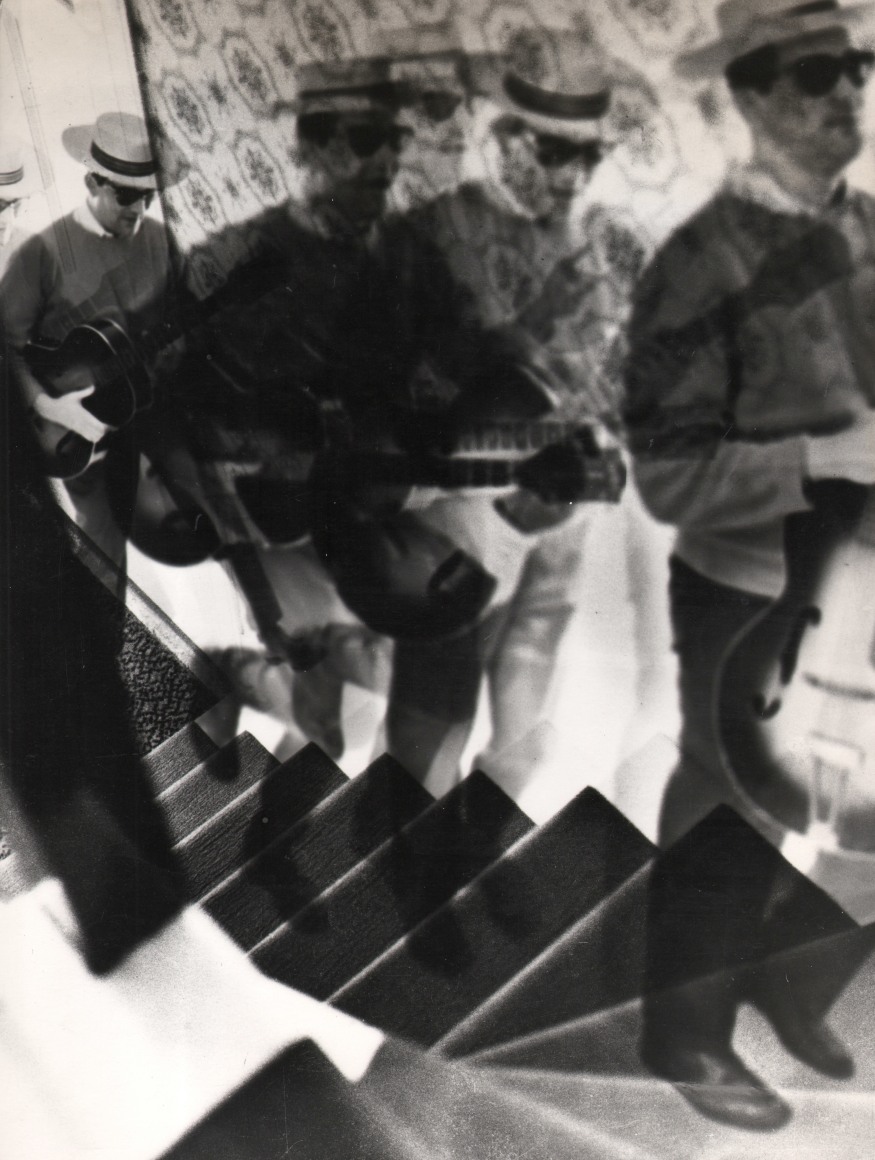 14. David Attie, Saloon Society, ​1959. Composite photograph featuring the same figure repeated up a flight of stairs. The figure is a man wearing a hat and sunglasses carrying a guitar.