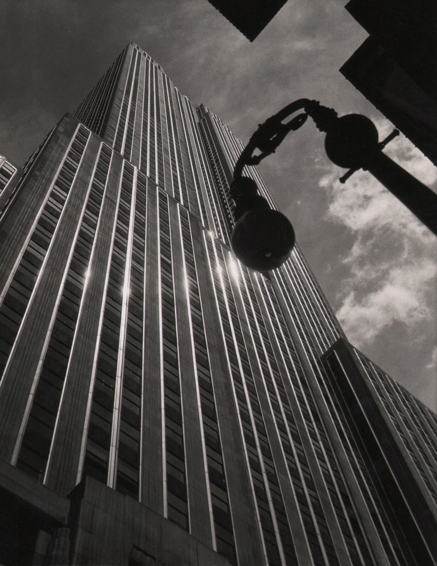 12. John C. Hatlem, Empire State Building, ​c. 1935. Upward-looking street-level view from below the reflective Empire State Building. A streetlight is silhouetted against a clouds and sky on the right of the frame.