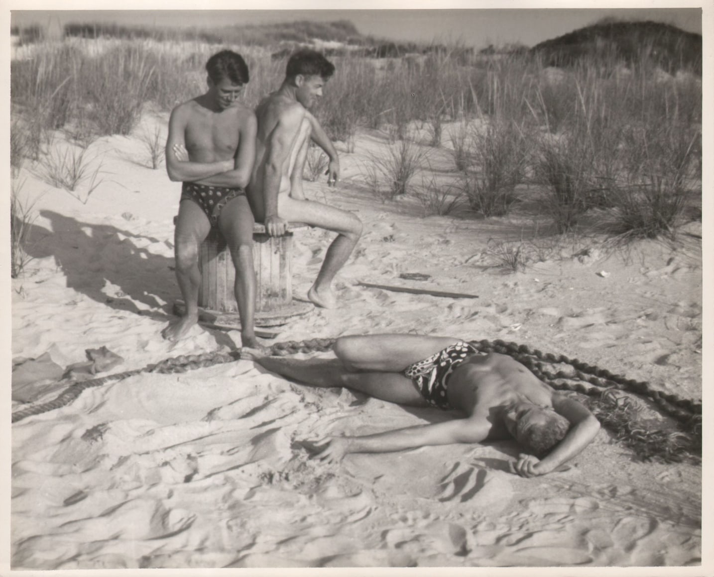 PaJaMa, Glenway Wescott, Paul Cadmus, George Platt Lynes, c. 1941. Two men sit on the beach on a large wooden spool (left), a third man lays in the sand, curving his body alongside a thick rope.
