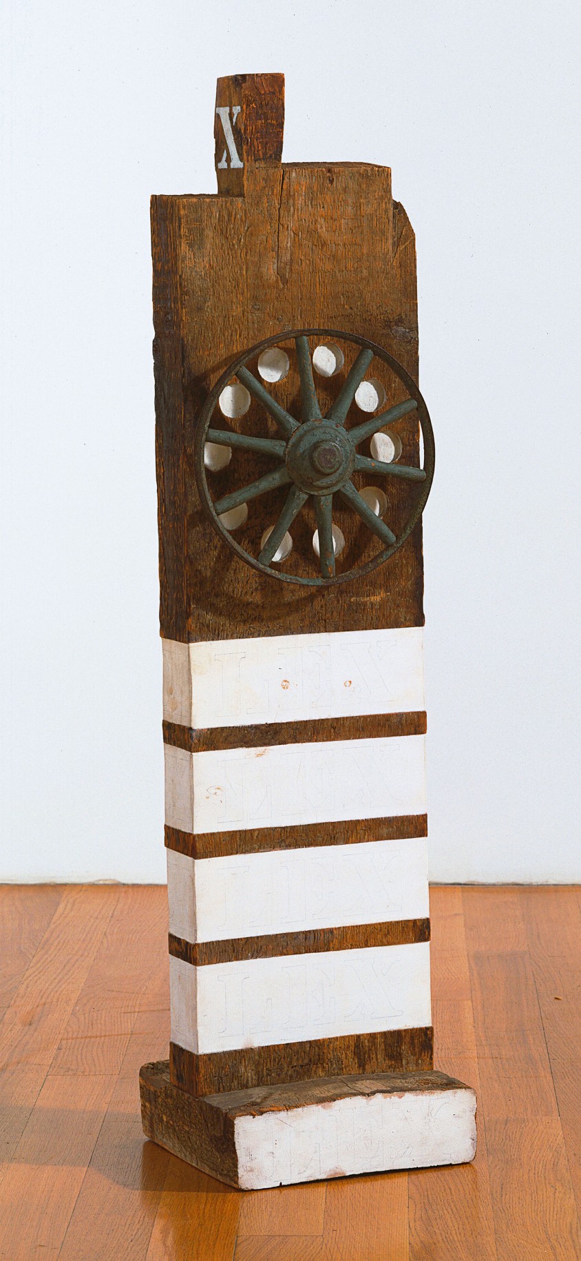 Law, a 44 1/8 by 10 7/8 by 3 1/8 inch sculpture consisting of a wooden beam with a haunched tenon on a wooden base. An iron wheel is affixed to the upper half of the front of the work; in between each spoke is a white painted orb. Four white stripes are painted around the bottom half of the sculpture, and the front of the base is also painted white.