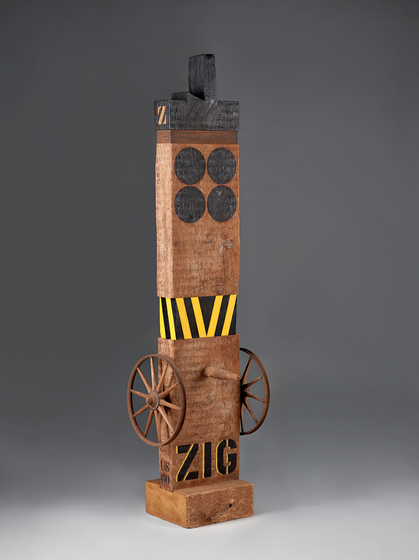 A 65 by 17 3/4 by 16 1/8 inch sculpture consisting of a wooden beam with a haunched tenon on a wooden base. Its title, &quot;Zig,&quot; is painted in black stenciled letters across the bottom of the work. Above the title, affixed on the left and right sides of the sculpture, are metal wheels. A wooden peg has been affixed to the front of the sculpture, in between the top rims of the wheels. A few inches above the peg is a band of yellow and black vertical stripes. The tenon and top of the sculpture have been painted black. Below this is a band of thin metal wires, and below that two rows of two black circles.