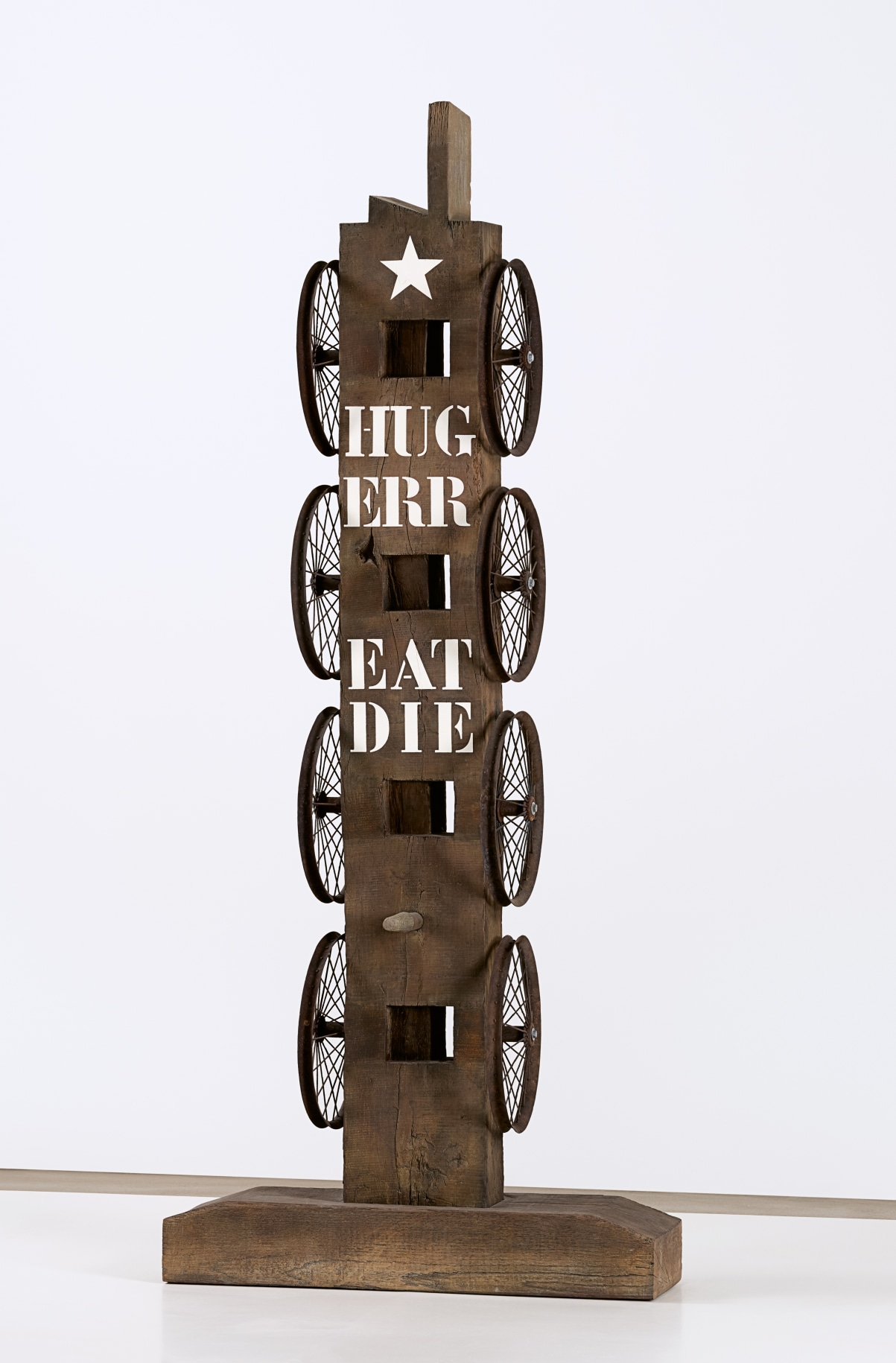 A 83 7/8 by 35 1/2 by 11 13/16 inch painted bronze sculpture consisting of a beam with a haunched tenon on a base. Four wheels run down the left and right sides of the sculpture. At the front top is a white star, below it the words &quot;Hug,&quot; &quot;Err,&quot; &quot;Eat,&quot; and &quot;Die&quot; are painted in white stenciled letters.