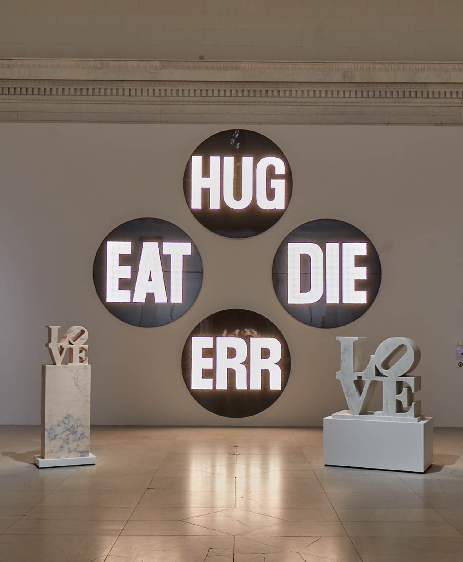 The Electric American Dream, a sculpture consisting of four 78 by 78 by 7 inch circular components. Each contains a different three letter word with lightbulbs that flash on and off. The words are eat, die, hug, and err.