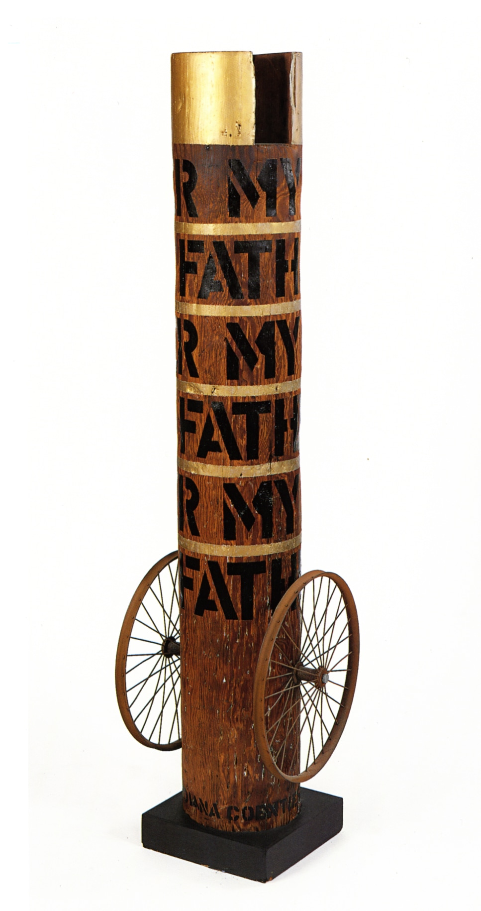 Column with its title &quot;My Father&quot; painted in five rows around the column in black stenciled letters. The top of the column is painted gold, and thin gold strips around the column separate each row of text. At the bottom of the column, on both sides, is a wheel.
