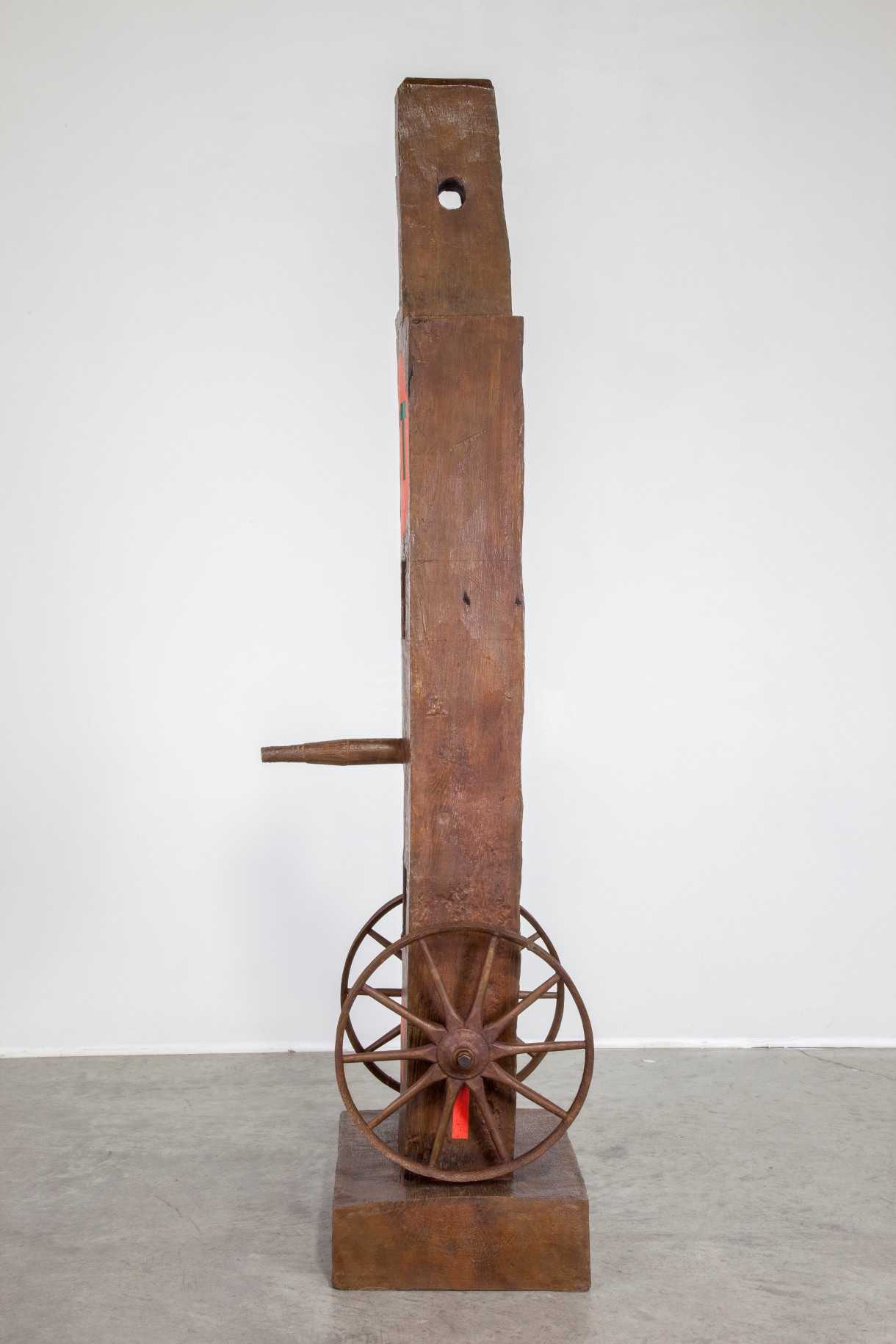 Right profile view of the bronze herm Eat. A red stenciled letter &quot;I&quot; is painted above the base. Above this a wheel has been attached. A whole in the wood is visible in the in the sculpture's haunched tenon.