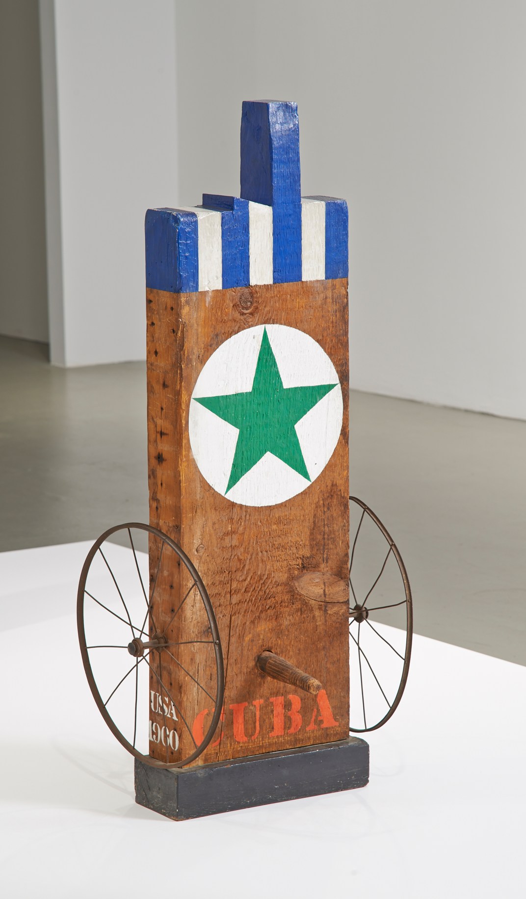 A 44 3/8 by 14 1/8 by 5 3/4 inch sculpture consisting of a wooden beam with a haunched tenon, standing on a wooden base. The work's title, Cuba, is painted in red letters across the bottom of the plank. Above it is a wooden peg, and to each side is an iron wheel. A green star in a white circle is found in the upper half of the sculpture, and above this the top part of the sculpture, including the tenon, is painted in blue and white vertical stripes.