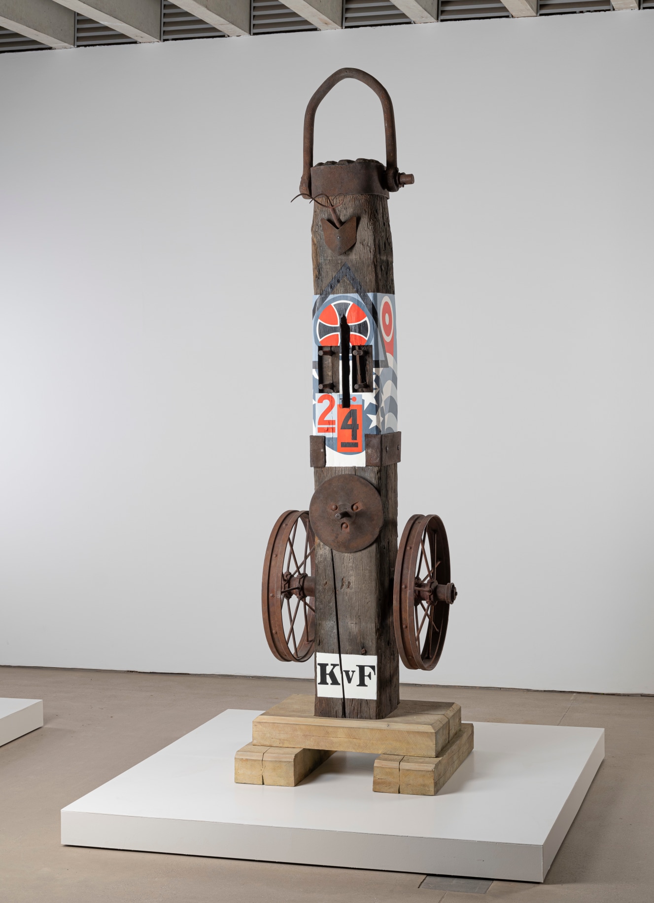 A 138 1/8 by 38 9/16 by 45 1/16 inch painted bronze sculpture consisting of a beam on a base. The work's title, &quot;KvF&quot; appears in stenciled letters against a white ground. A wheel is affixed to the right and left sides of the sculpture. On the front of the sculpture, level with the top of the wheels, is a disk. Above this a section of the sculpture has been painted with red, blue, white, and black designs that include a cross, stars, and the numbers 2 and 4. At the top of the sculpture is a cap with a handle.