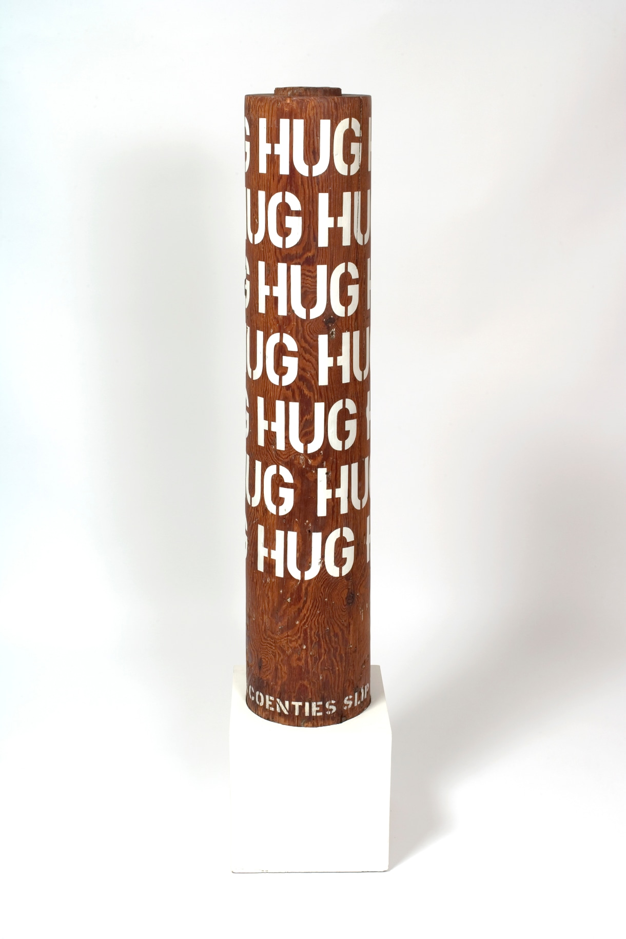 A column with seven rows of the word hug in gesso stenciled letters wrapping around the upper three quarters of the sculpture.