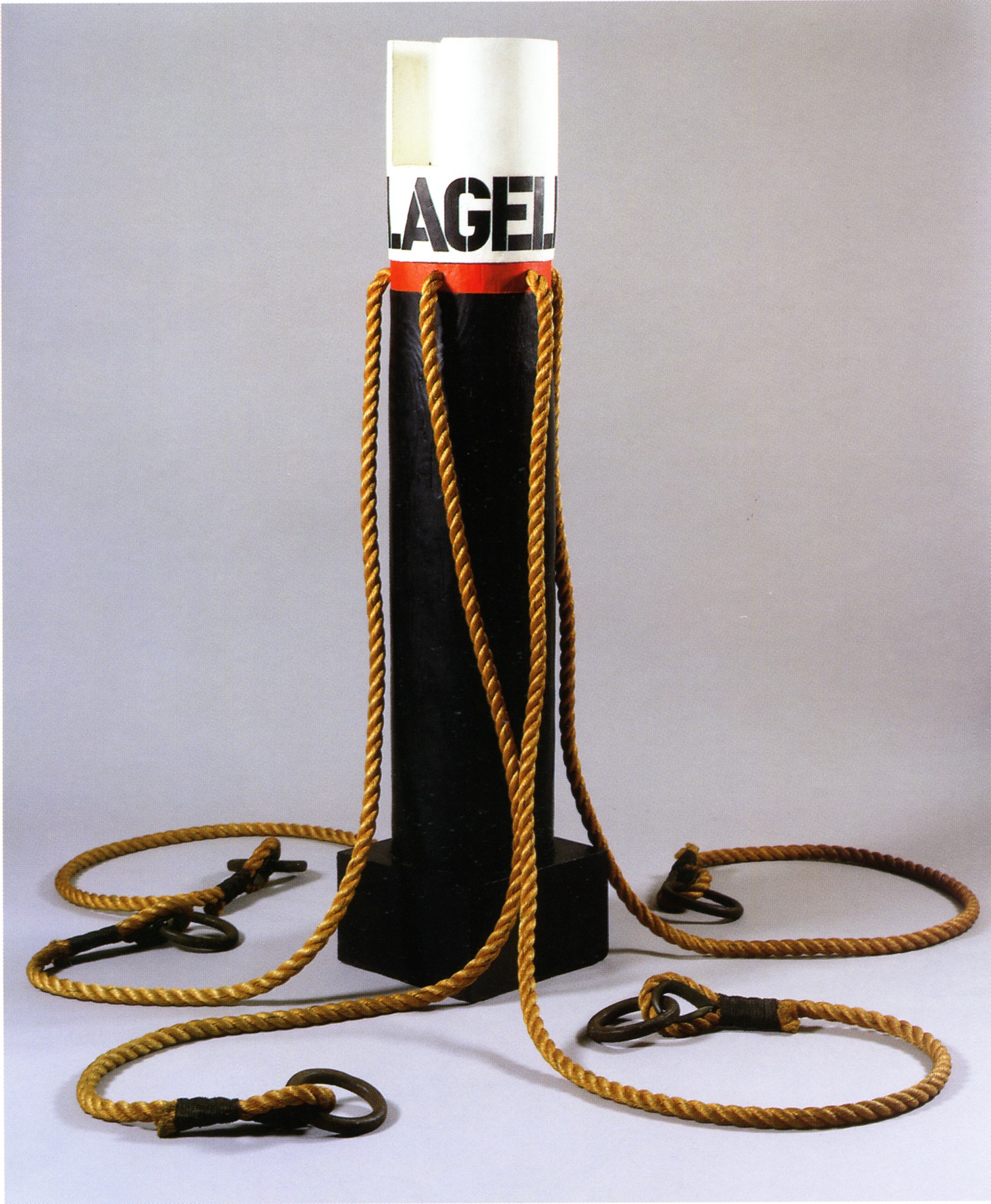A 63 1/3 inch high wooden column on a wooden base. The top of the column has been painted white, with the work's title, Flagellant, painted in black stenciled letters. Below the title is a red band, where four long ropes with shackles have been attached to the work.