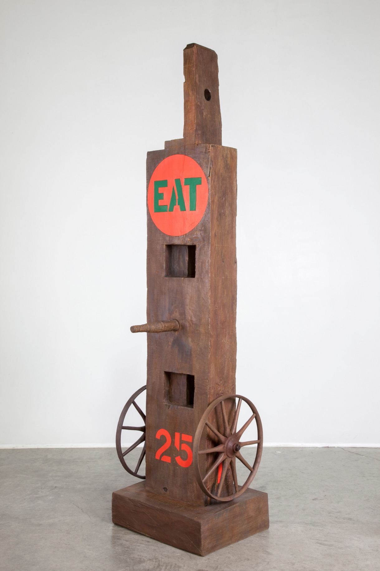 Eat is a 58 5/8 by 15 by 12 5/8 painted bronze sculpture of a beam with a tenon, standing on a base. A wheel is affixed to the right and left sides at the bottom of the sculpture. In between a red number 25 has been painted. A peg protrudes from the center of the sculpture, and at the top of the sculpture is a red circle containing the word &quot;eat&quot; painted in green stenciled letters.