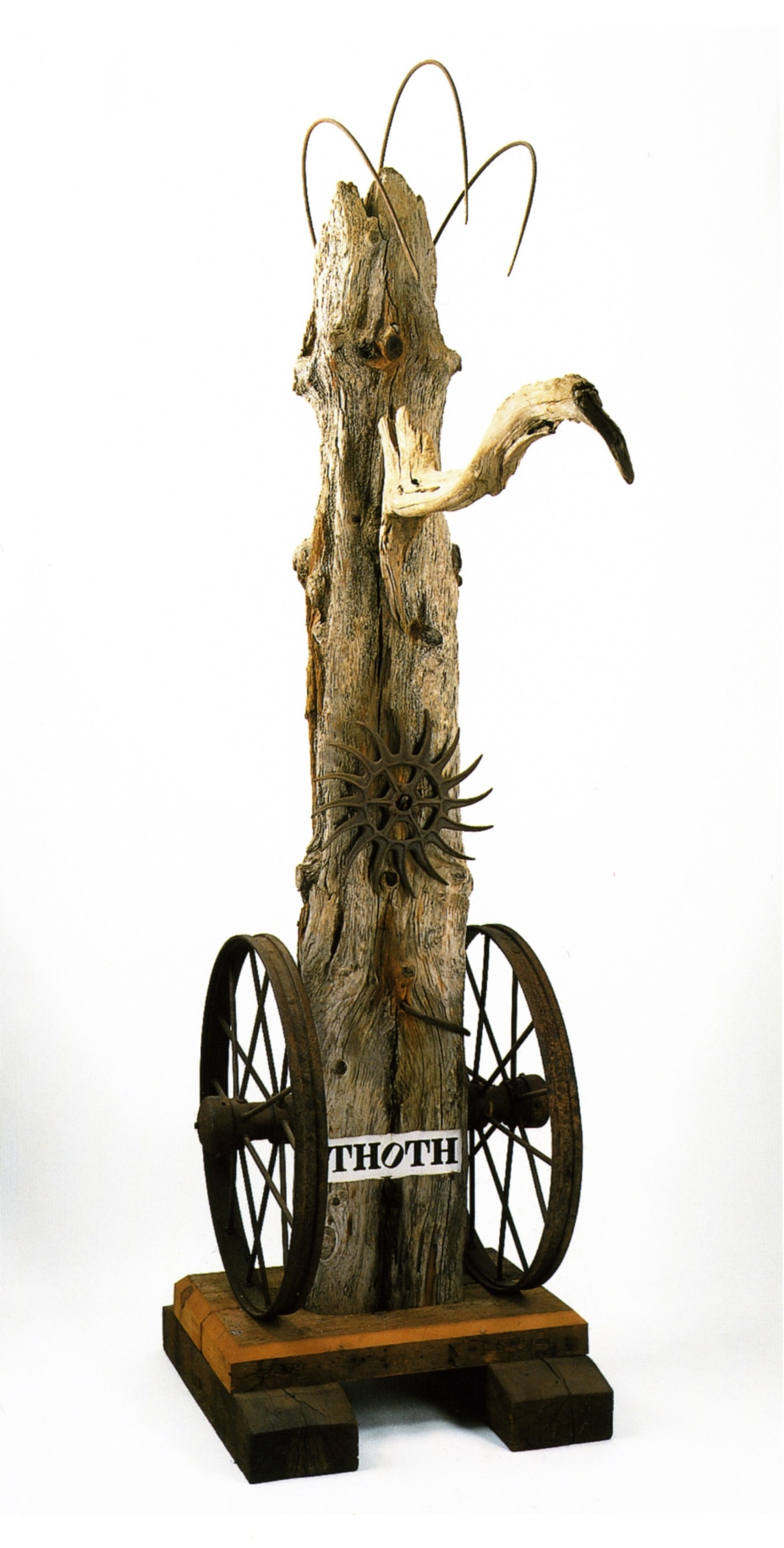 A 130 1/2 by 71 by 38 1/2 inch weathered wood sculpture on a wooden base. The sculpture's title, &quot;Thoth,&quot; is painted in black stenciled letters on a white ground across the front of the sculpture, near the base. An iron wheel has been attached to the sides of the sculpture to the left and right of the title. A long metal nail protrudes from the sculpture above the title, and above this is an old rotary tiller wheel. A piece of driftwood, resembling a long beak, has been attached to the front of the work near the top. to