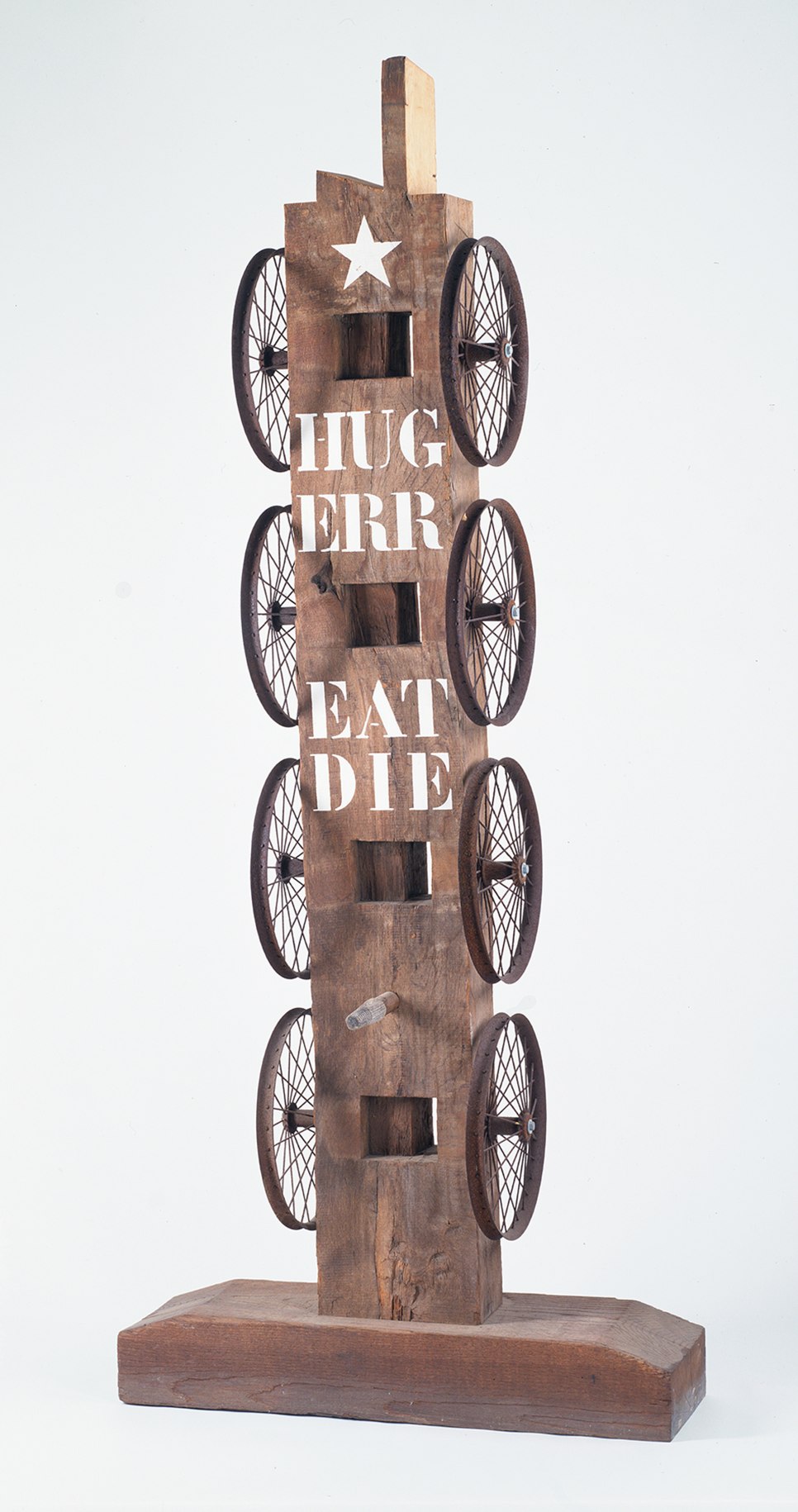 A 84 by 35 1/2 by 17 inch sculpture consisting of a wooden beam with a haunched tenon on a wooden base. Four iron wheels run down the left and right sides of the sculpture. At the front top is a white star, below it the words &quot;Hug,&quot; &quot;Err,&quot; &quot;Eat,&quot; and &quot;Die&quot; are painted in white stenciled letters.