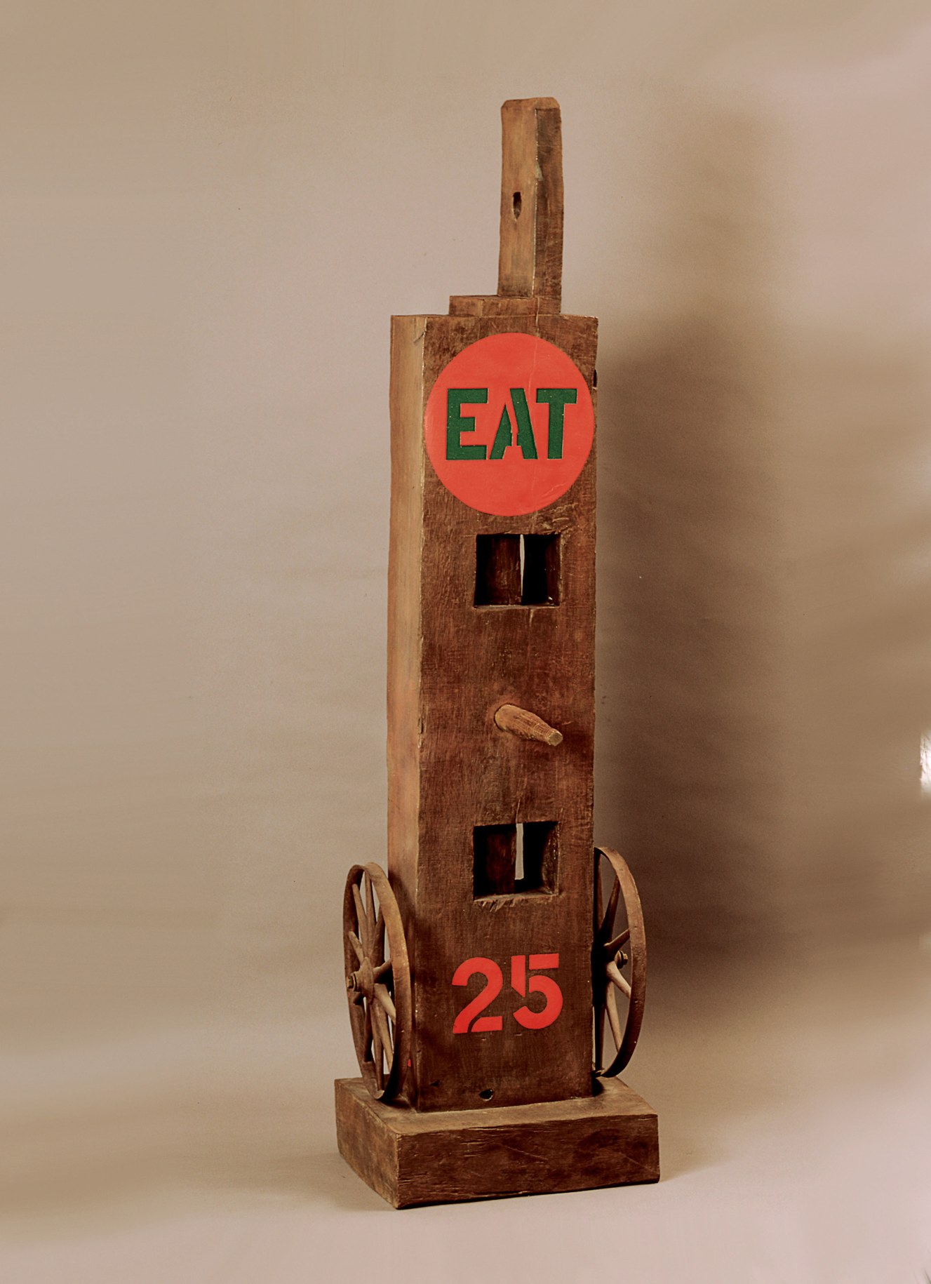 Eat, a 60 by 15 1/2 by 17 inch sculpture consisting of a wooden beam with a haunched tenon, standing on a wooden base. An iron and wooden wheel is affixed to the right and left sides at the bottom of the sculpture. In between a red number 25 has been painted. A wooden peg protrudes from the center of the sculpture, and at the top of the sculpture is a red circle containing the word &quot;eat&quot; painted in green stenciled letters.