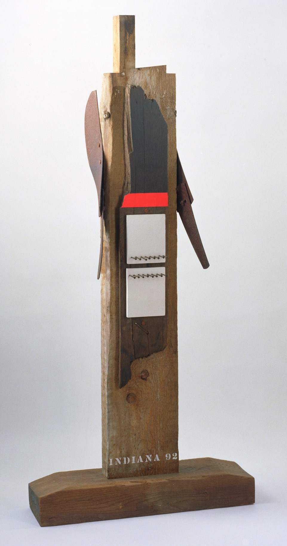 The verso of the sculpture Hero. It is signed and dated &quot;Indiana 92&quot; in white stenciled letters across the bottom of the beam. A long, thin piece of wood has been affixed to the center. It is been painted with a red band and two white rectangular pieces of wood, each with a row of nails, have been affixed to it.
